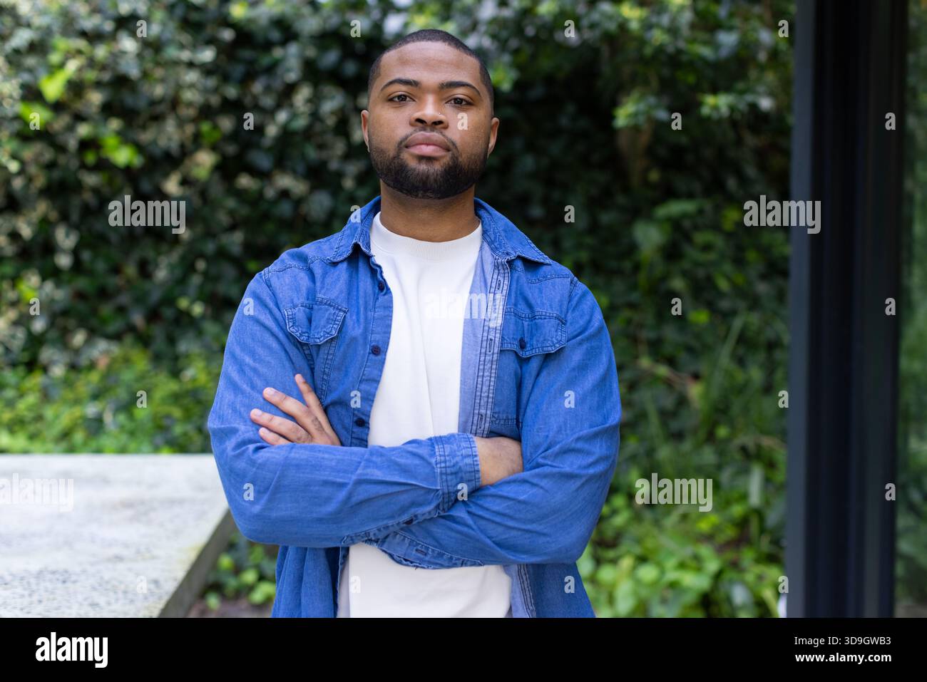 Uomo afroamericano in piedi con le braccia incrociate e indossa una camicia in denim su una panchina di cemento in mezzo al muro di edera Foto Stock