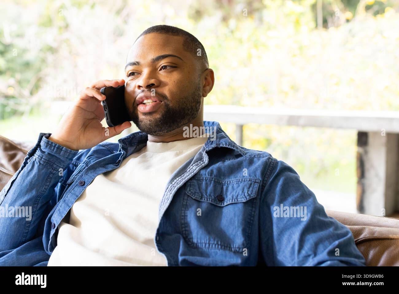 Uomo afroamericano seduto su un divano in pelle a casa sotto una grande finestra che parla sullo smartphone Foto Stock