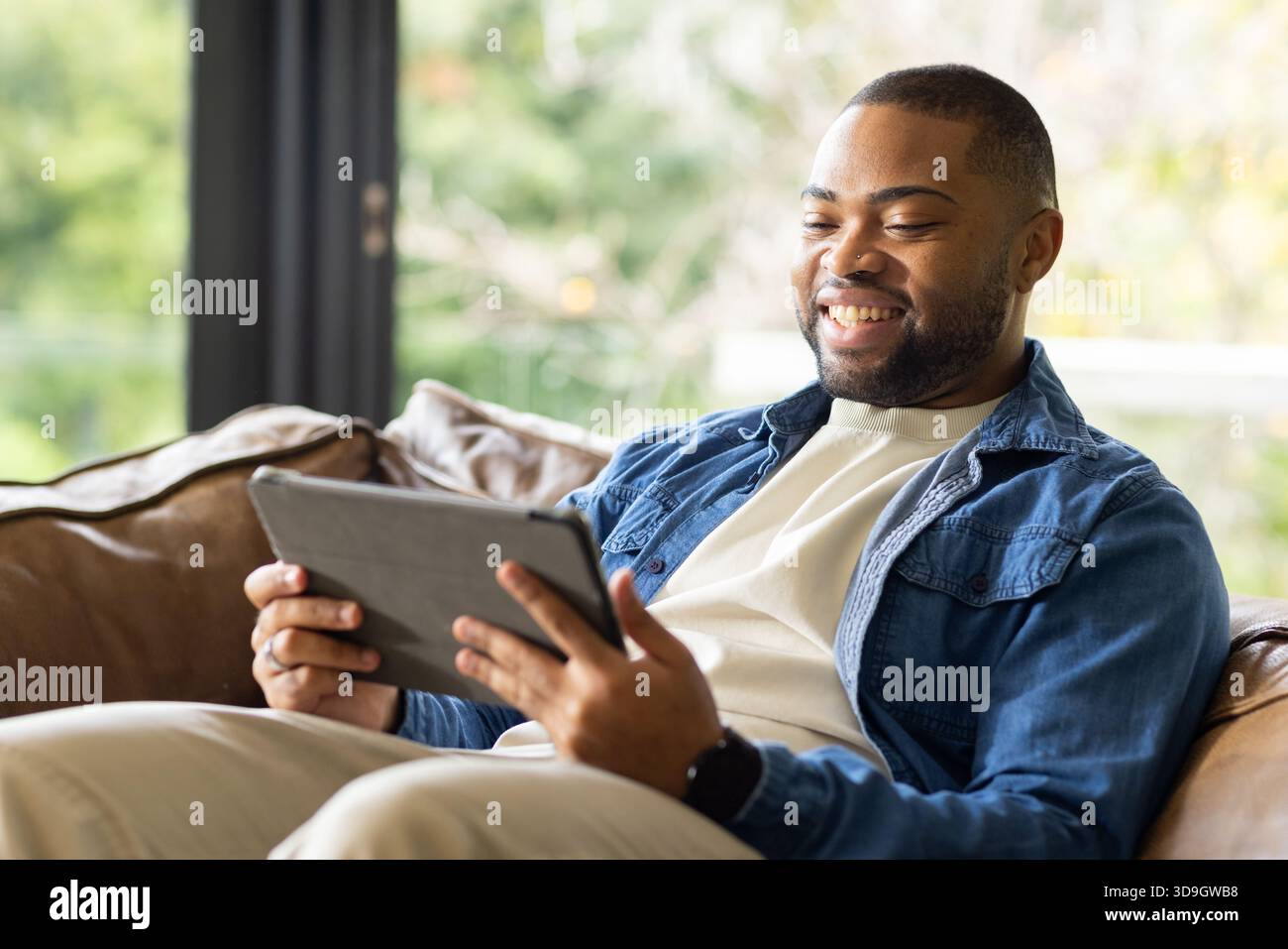 Uomo afro-americano sorridente, tenendo un tablet seduto sul divano vicino alla finestra in soggiorno, spazio fotocopie Foto Stock
