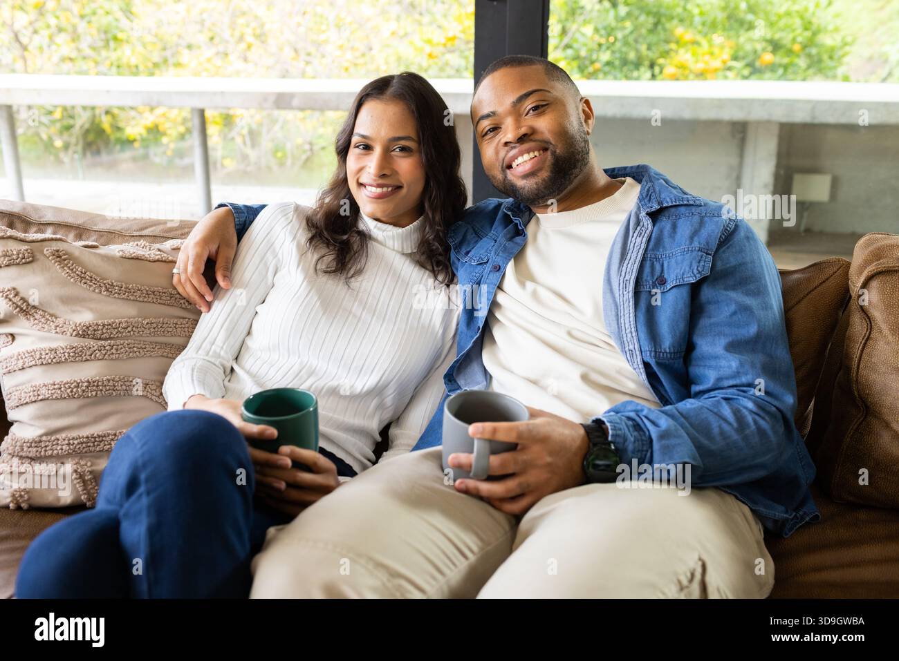 Diverse coppie che tengono tazze in ceramica mentre si siedono sul divano marrone accanto alla finestra e si affacciano sul giardino Foto Stock