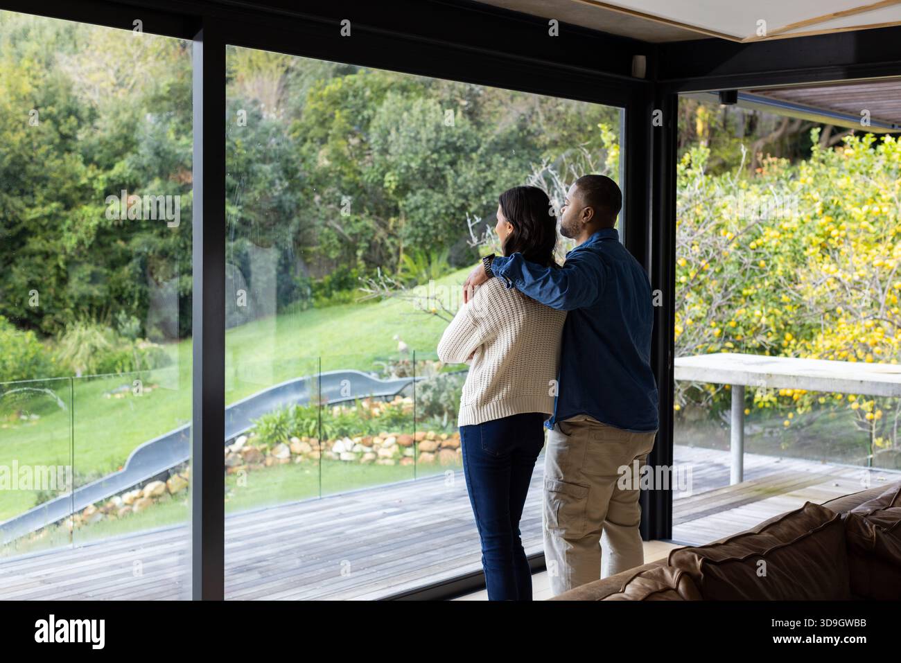 Coppia afroamericana in piedi nel soggiorno che guarda attraverso le pareti di vetro sul ponte di legno e lo scivolo Foto Stock