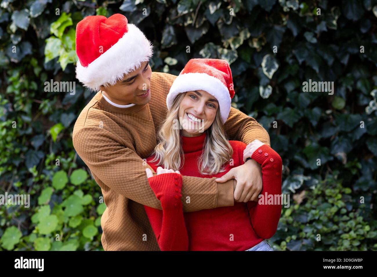 Coppie diverse che si abbracciano e sorridono indossando cappelli di Babbo Natale e maglioni in maglia in giardino Foto Stock