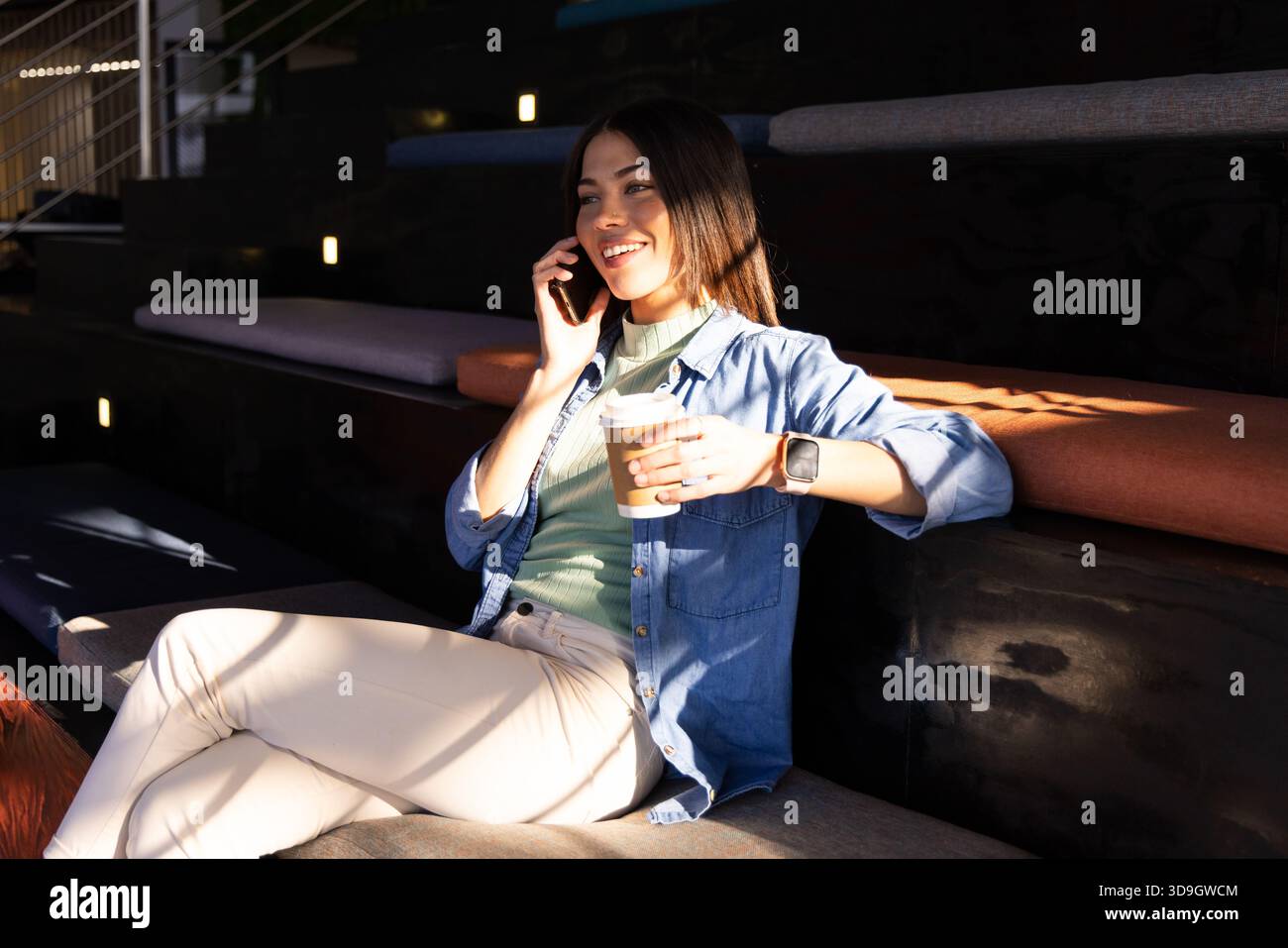 Donna indiana che tiene lo smartphone all'orecchio, indossa uno smartwatch, sorseggia una tazza di caffè in salotto Foto Stock