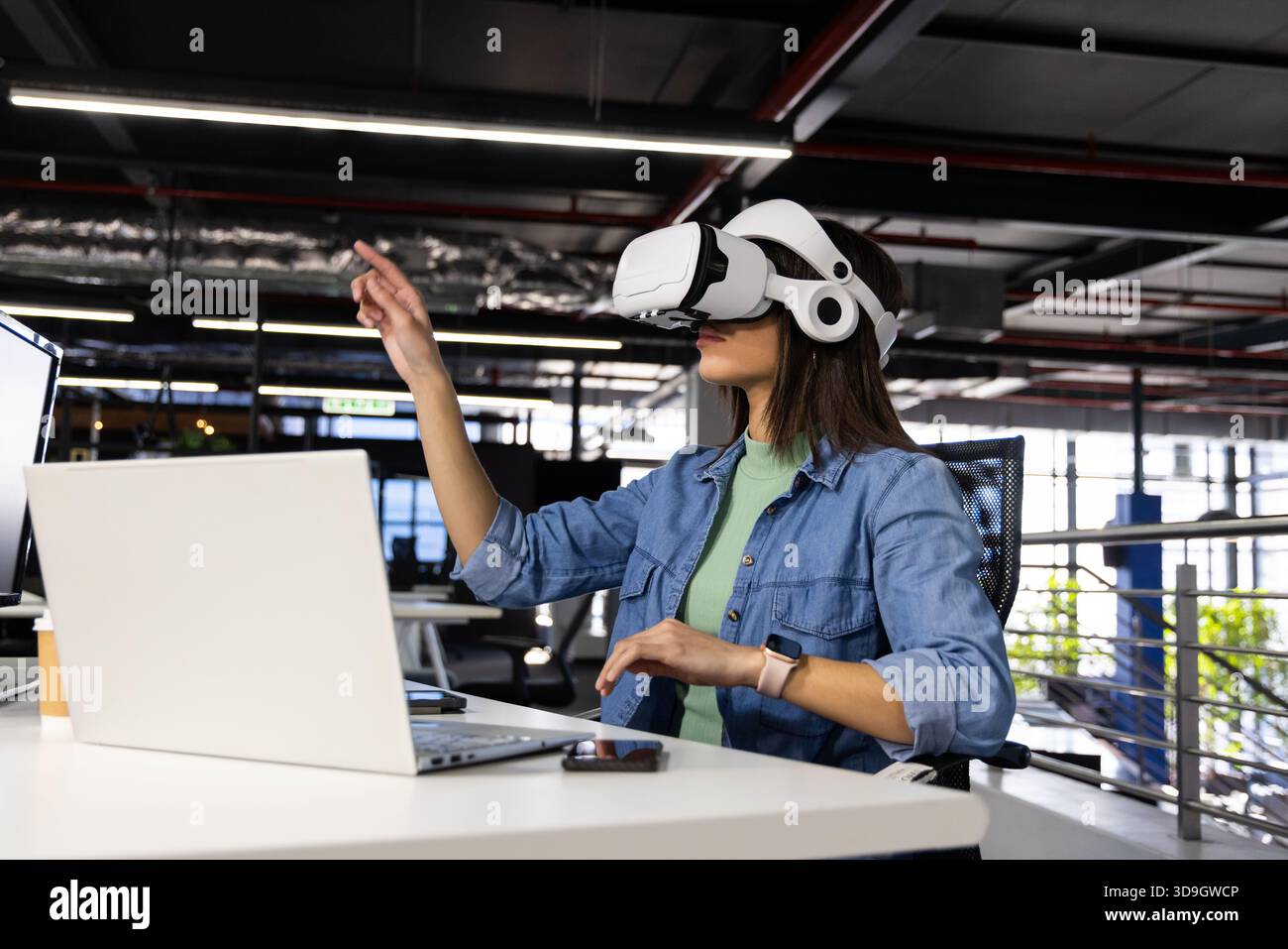 Laptop e smartphone appoggiati su una scrivania da ufficio bianca con illuminazione LED lineare, visore VR, tazza da caffè Foto Stock