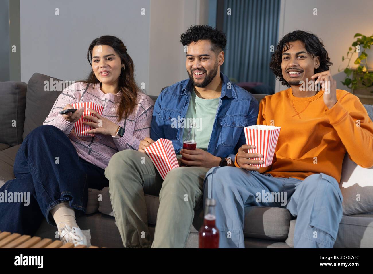 Diversi amici seduti su un divano grigio nel soggiorno che regala popcorn e bibita rossa sorseggiando a distanza Foto Stock