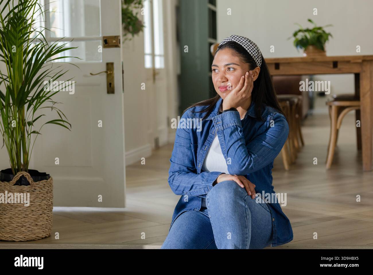 Donna che indossa una camicia in denim seduta sul pavimento di casa accanto alla porta di vetro aperta e alla pianta in vaso Foto Stock