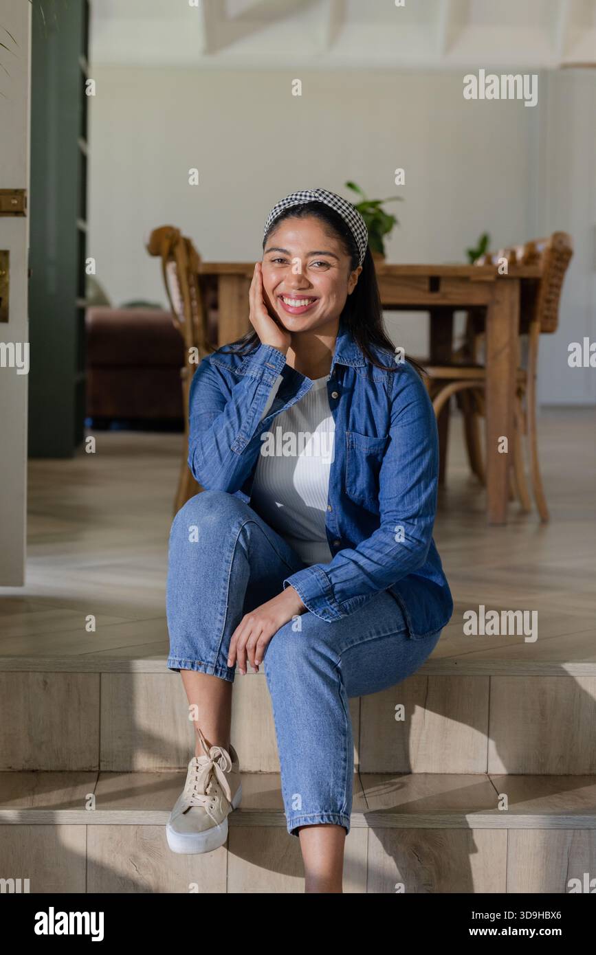Donna seduta su gradini di legno in una casa moderna sorridente e indossa camicia in denim e fascia a scacchi Foto Stock