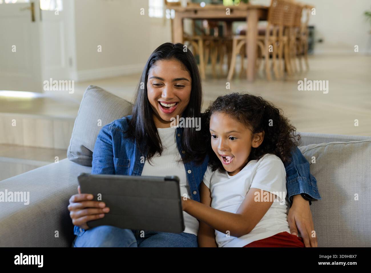 Madre asiatica e figlia afro-americana che tiene in mano una tavoletta nera mentre si siede sul divano grigio chiaro Foto Stock