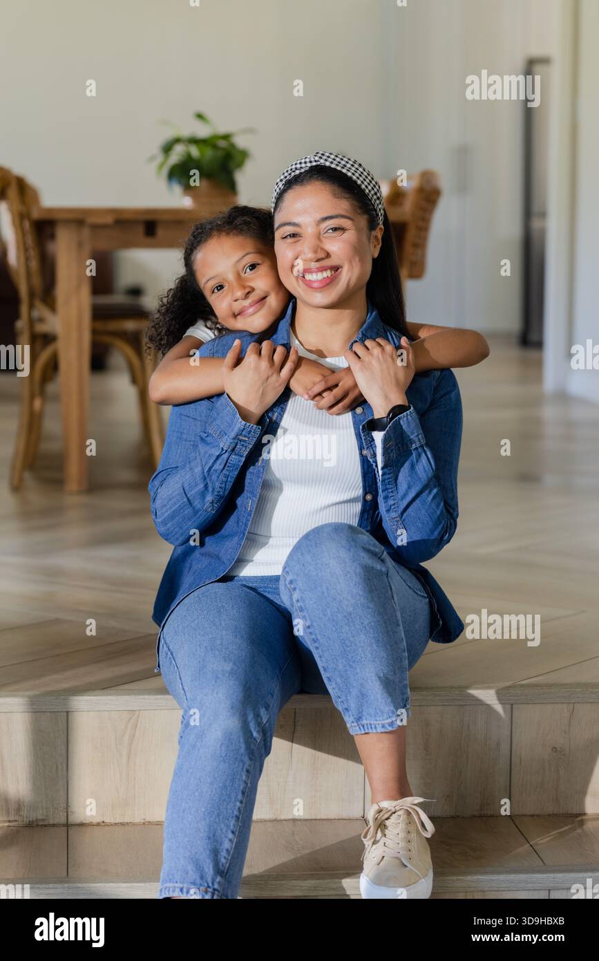 Madre e figlia si abbracciano su un gradino di legno nella zona pranzo del soggiorno di casa indossando abiti in denim Foto Stock