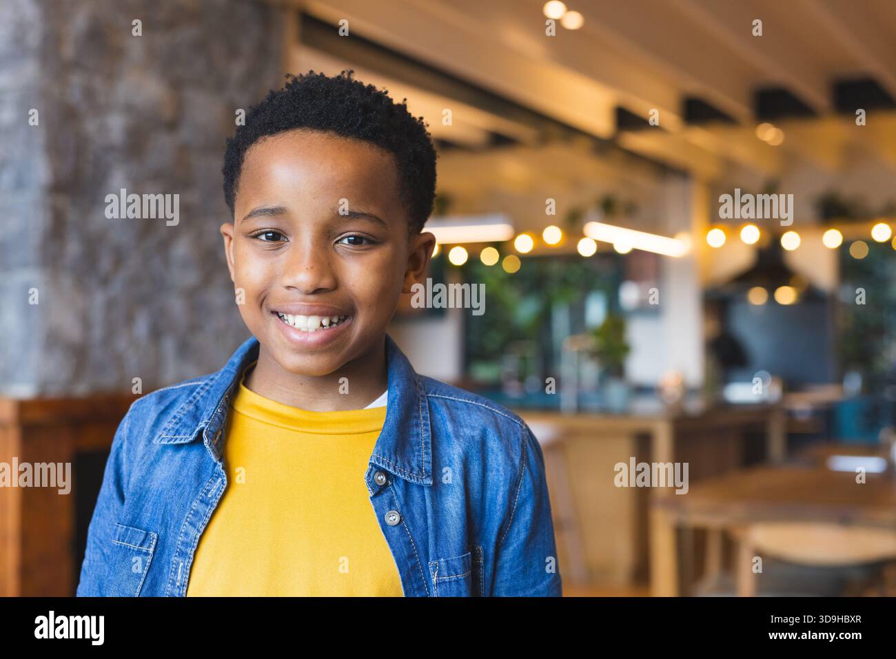 Sorridente ragazzo afroamericano in piedi all'interno del caffè con pareti in pietra, tavoli e luci a corda Foto Stock