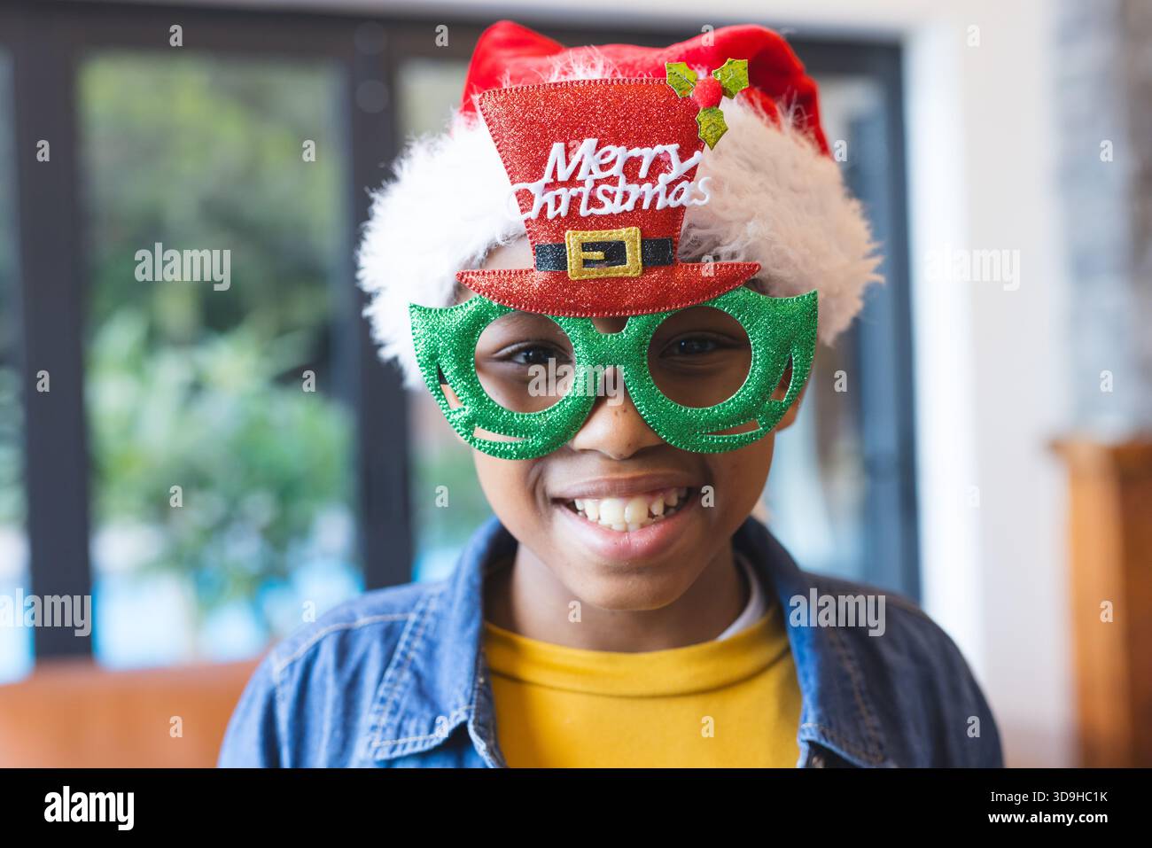 Ragazzo che indossa un buon cappello natalizio, occhiali verdi con glitter, giacca in denim e camicia gialla vicino alla finestra Foto Stock