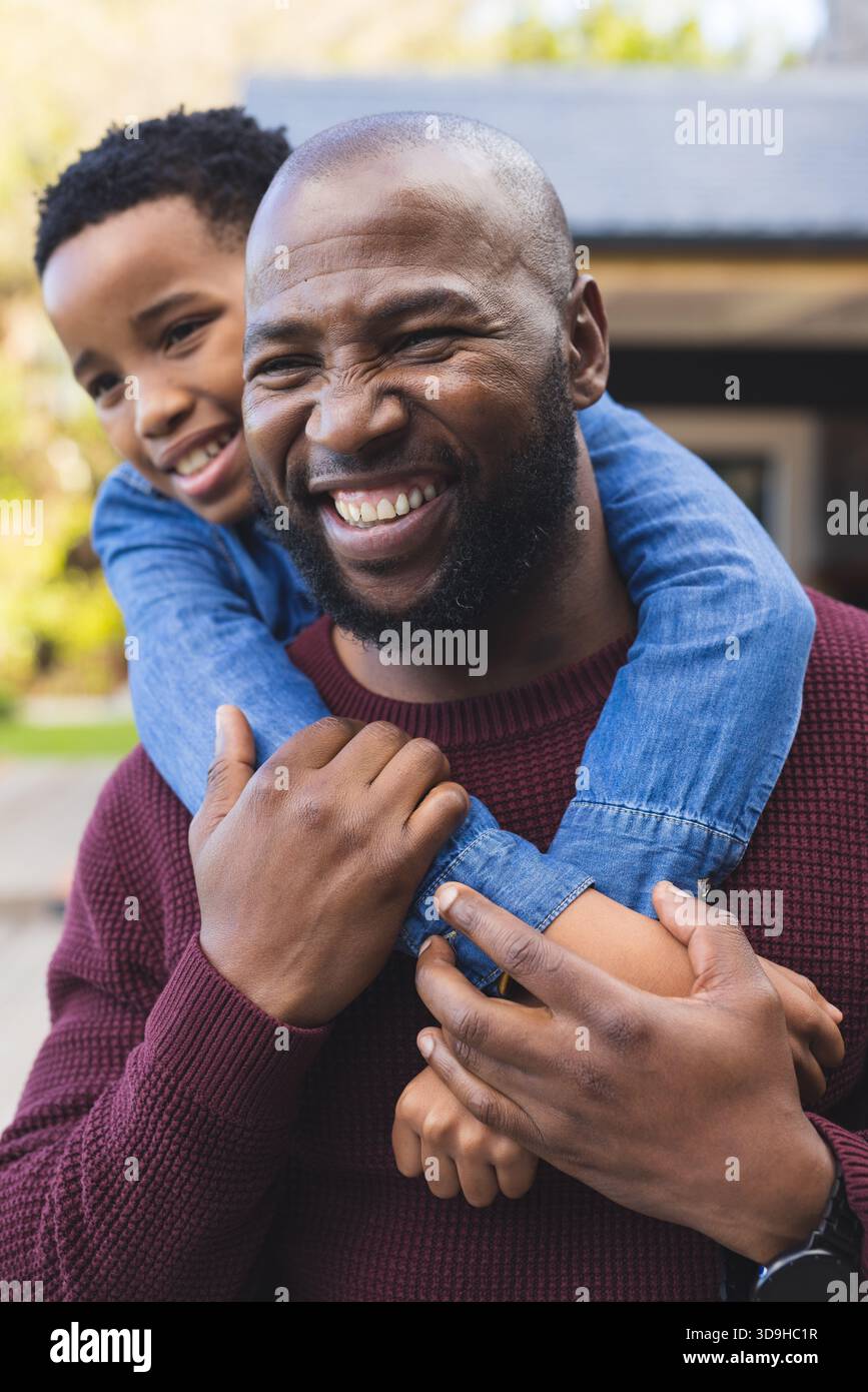 Padre afroamericano che porta il figlio che indossa una camicia blu sul retro di casa con smartwatch Foto Stock