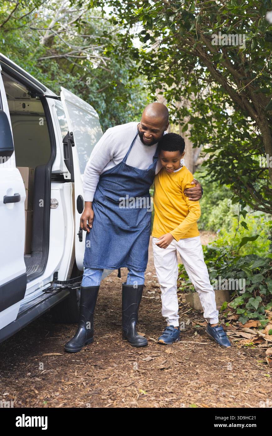 Padre e bambino afroamericano che indossano grembiule appoggiato al furgone bianco sul vialetto che tiene le chiavi Foto Stock