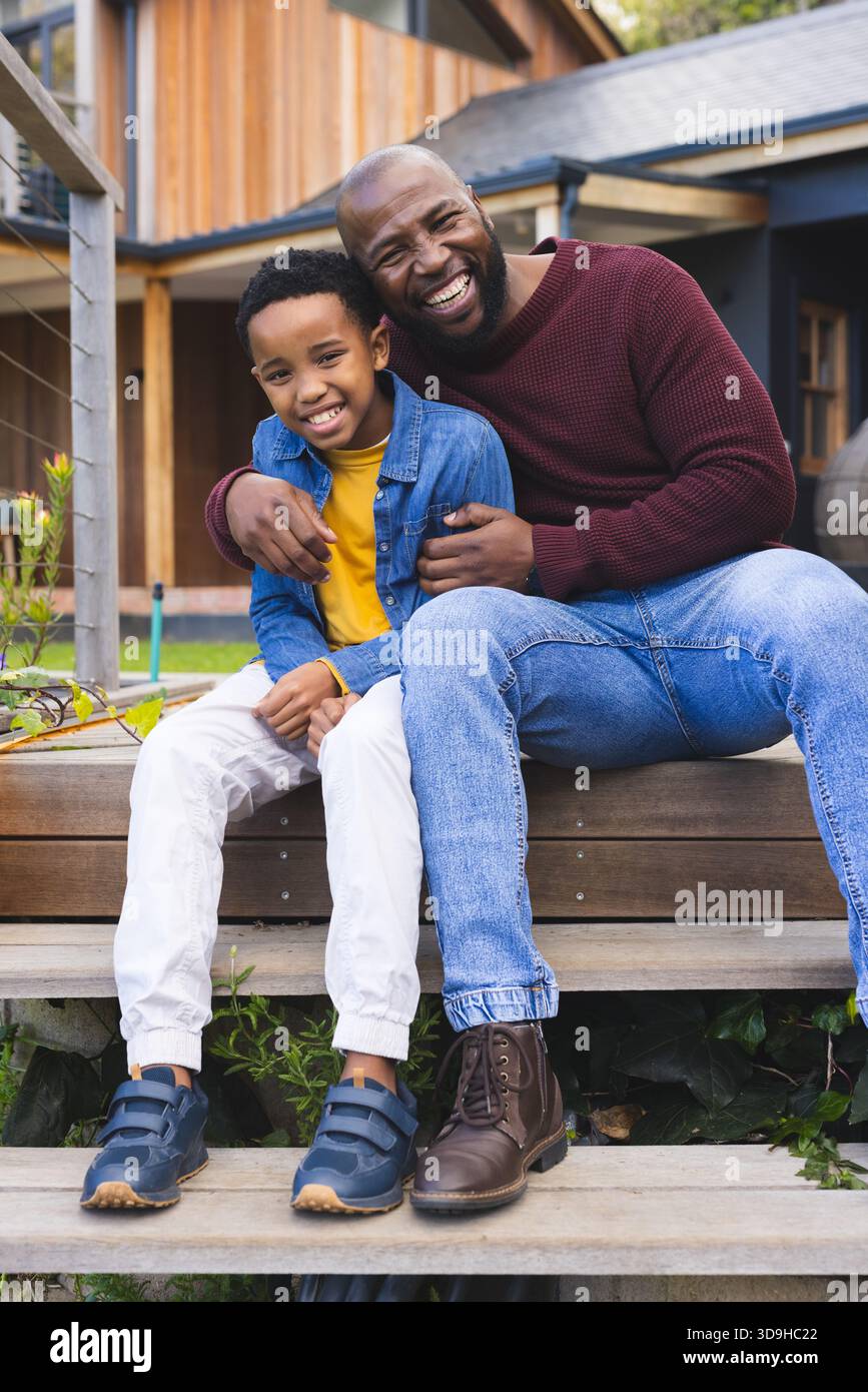 Padre e figlio afroamericani che abbracciano e sorridono su gradini di legno prima del letto da giardino con il tubo Foto Stock