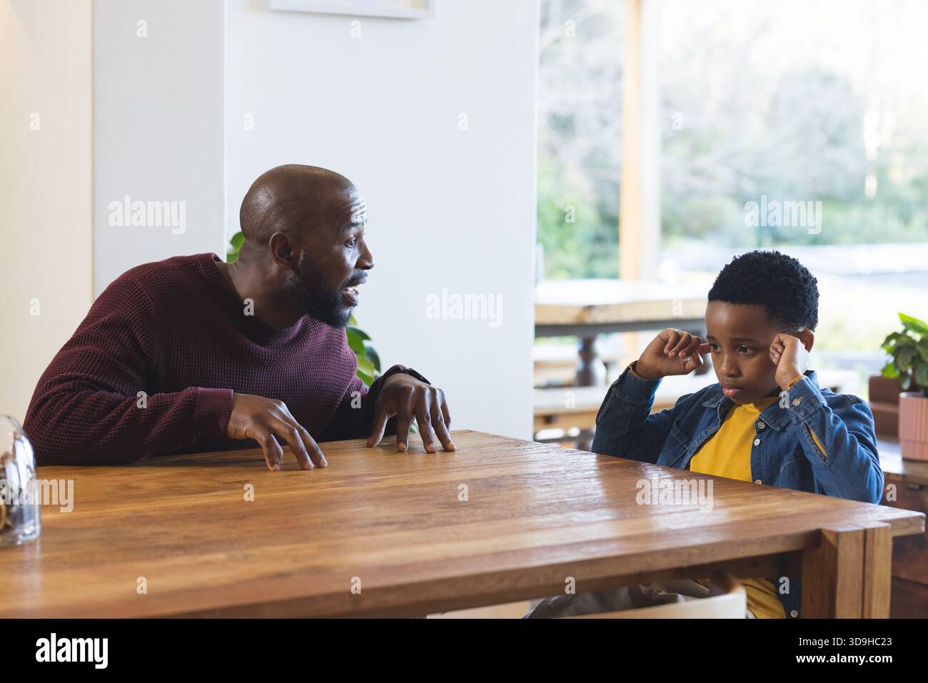 Padre e figlio afroamericani che parlano al tavolo da pranzo con cloche, piante mentre il ragazzo si toglie le orecchie Foto Stock