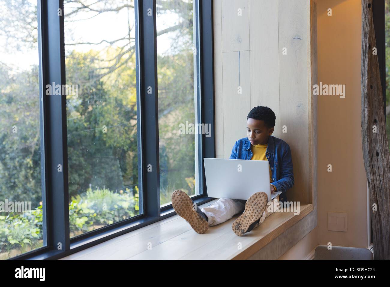 Bambino afroamericano seduto su una panchina di legno che studia su un computer portatile vicino alla finestra a griglia, copia spazio Foto Stock