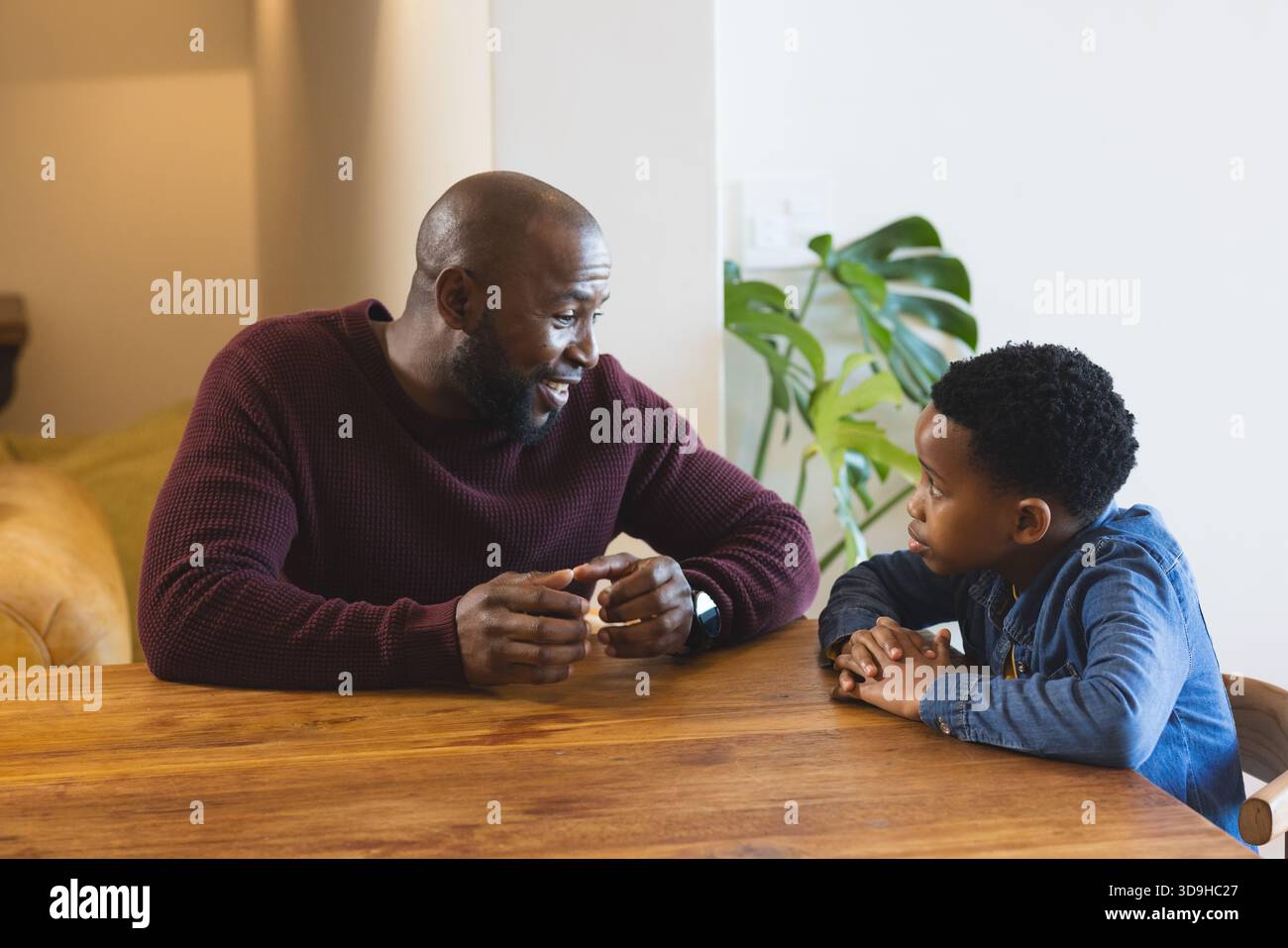 Padre e figlio afroamericani in maglione e camicia in denim impegnati in conversazione al tavolo di legno Foto Stock
