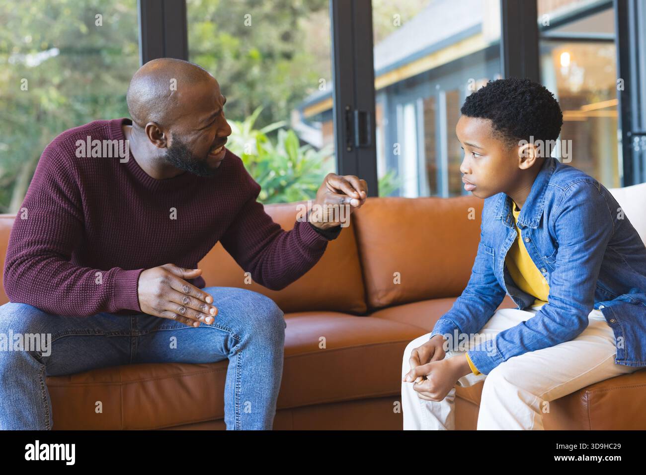 Padre afroamericano e figlio preadolescente che parlano, gestiscono sul divano in pelle marrone nel soggiorno Foto Stock
