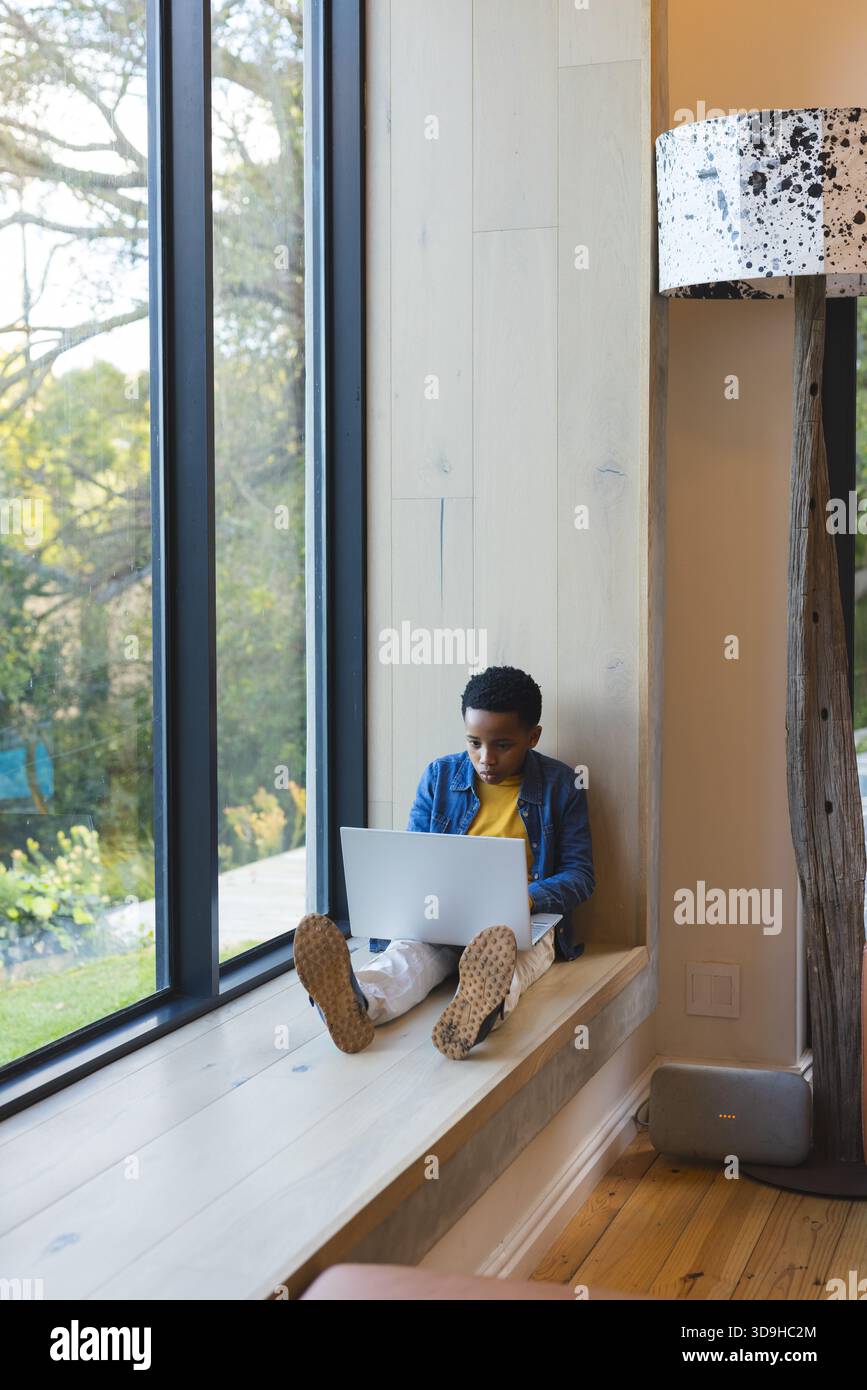 Bambino afroamericano maschio seduto su una panchina di legno della finestra usando il computer portatile accanto alla lampada da terra a casa Foto Stock
