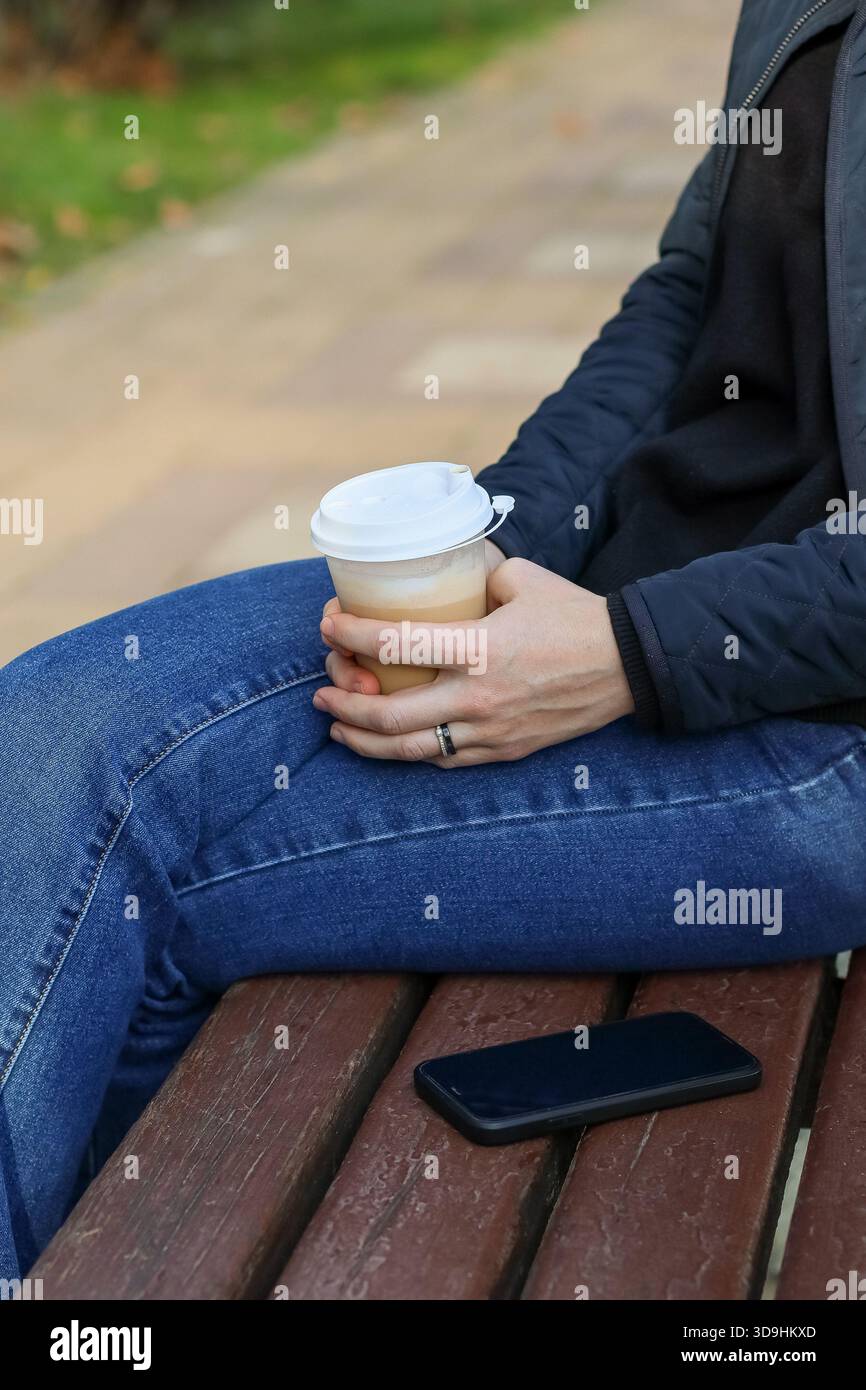 Individuale in jeans e giacca con caffè cremoso su panca di legno e telefono, circondato da un'area verde del parco. Foto Stock