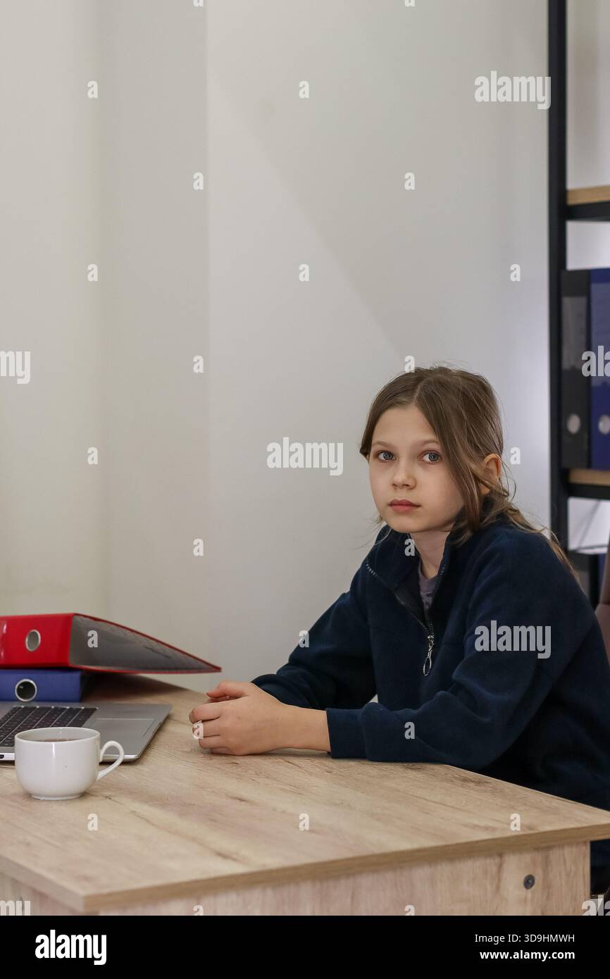 Una donna preadolescente dal look pensivo si siede al tavolo di legno, si mette le mani insieme vicino al computer portatile, circondata da leganti colorati e una tazza calda. Foto Stock