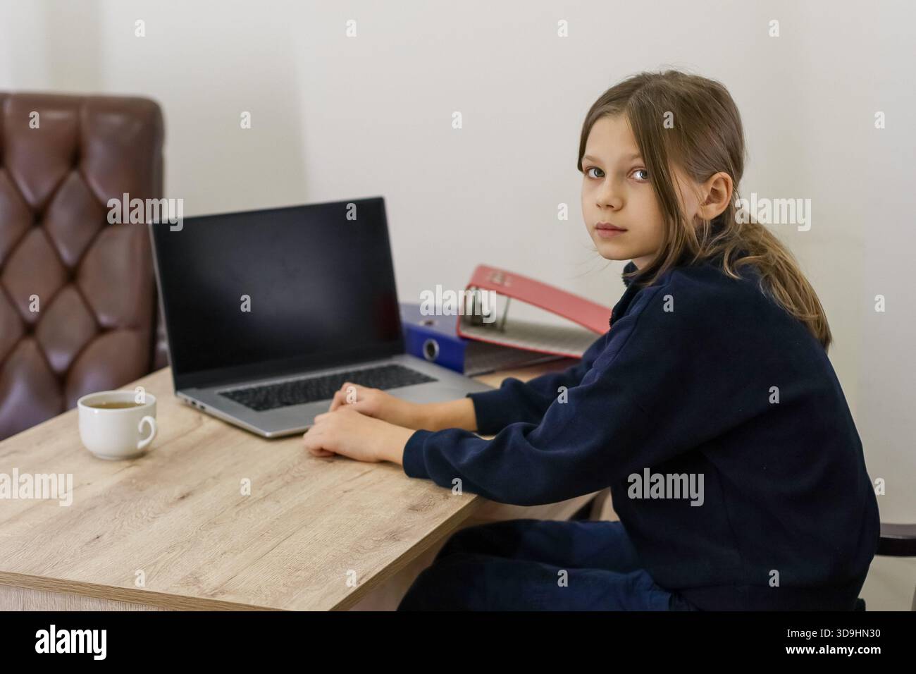 Una ragazza preadolescente in maglione blu siede sulla scrivania di legno con laptop spento, raccoglitori colorati e una tazza di tè, guardando da parte con attenzione. Foto Stock