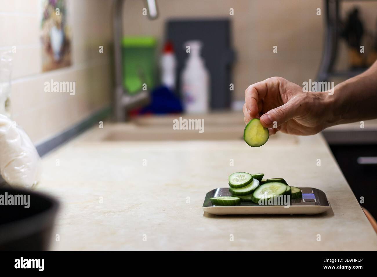 Foto dettagliata di dita che lasciano cadere una fetta di cetriolo su una pila di fette su una bilancia digitale da cucina, con elementi di sfondo da cucina. Foto Stock