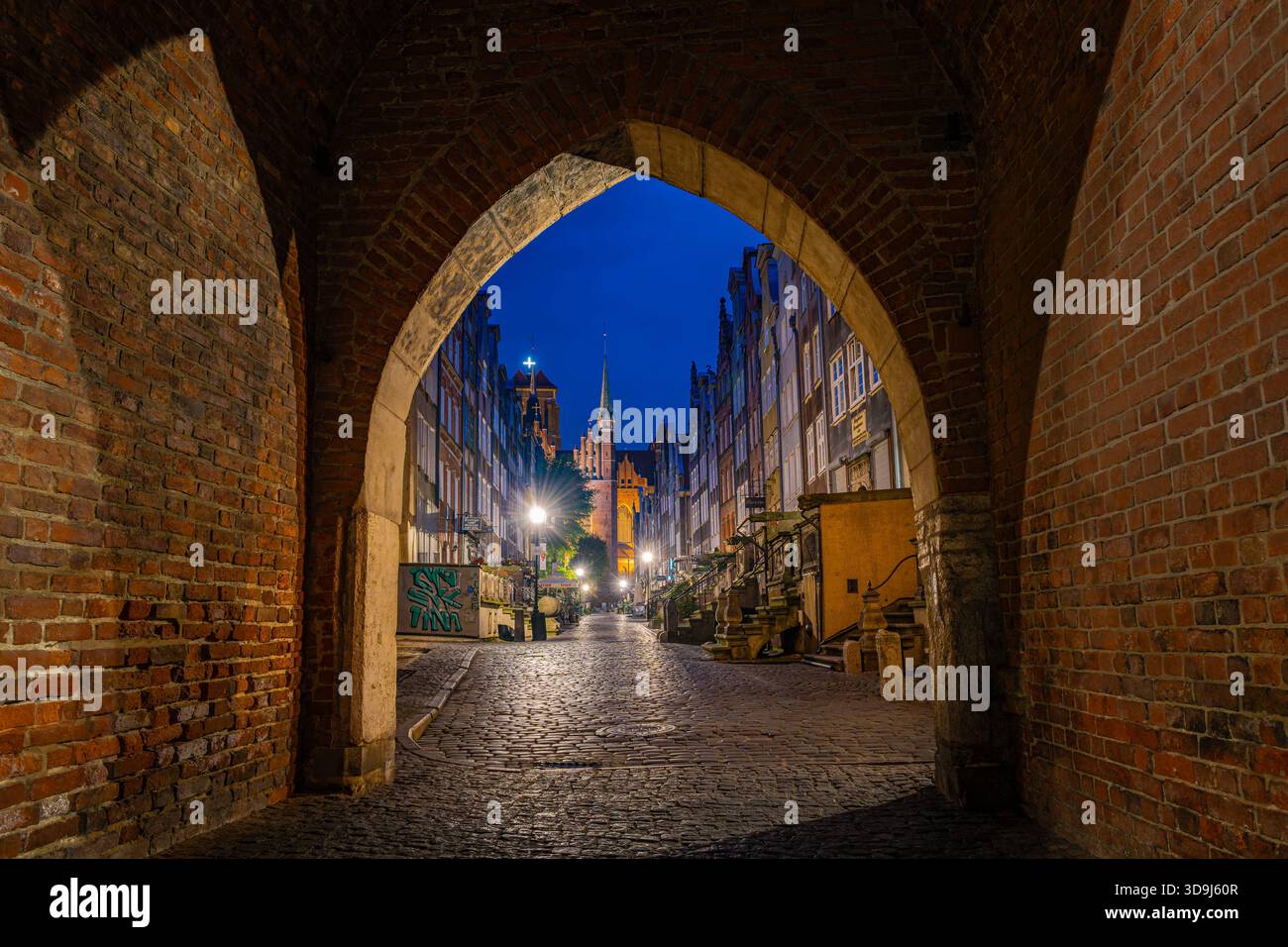 Arco medievale in mattoni che incornicia la strada acciottolata della città vecchia illuminata di Danzica all'ora blu, Polonia Foto Stock