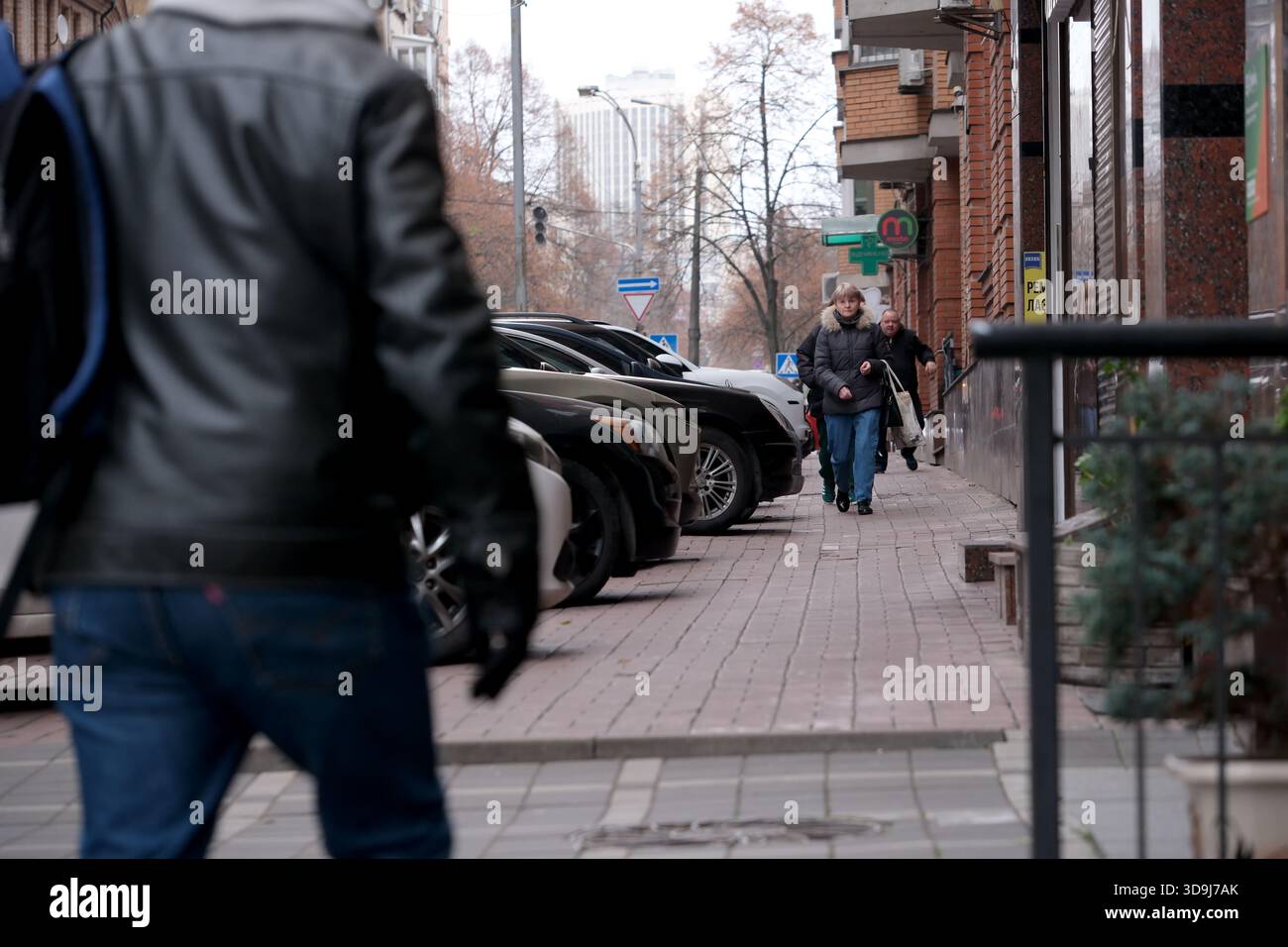 Due persone camminano accanto alle auto parcheggiate su un vivace marciapiede urbano. Kiev, Ucraina. 5 dicembre 2025. Foto Stock