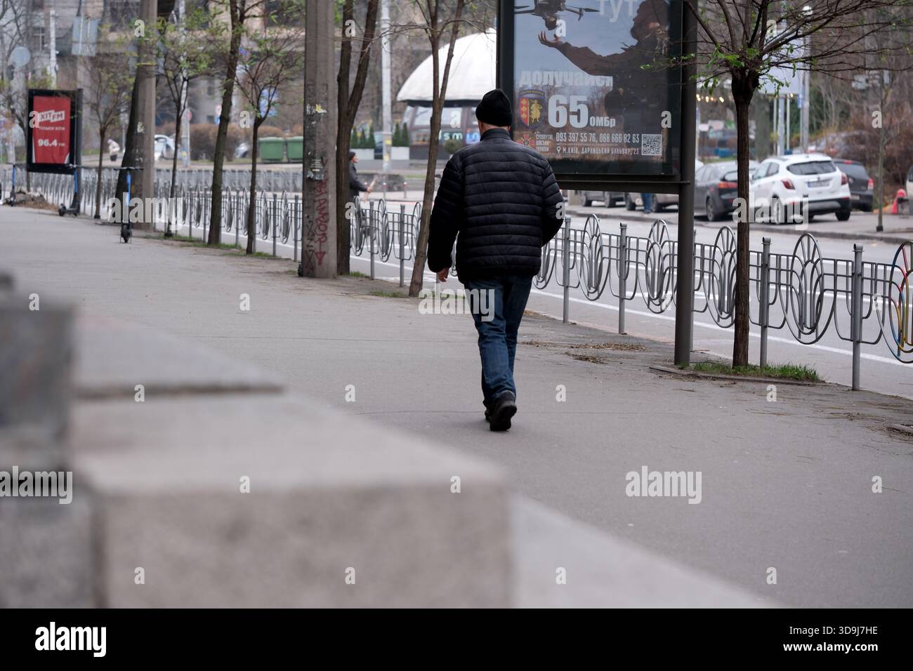 Una figura solitaria passeggia lungo una strada trafficata, circondata da alberi in erba. Kiev, Ucraina. 5 dicembre 2025. Foto Stock