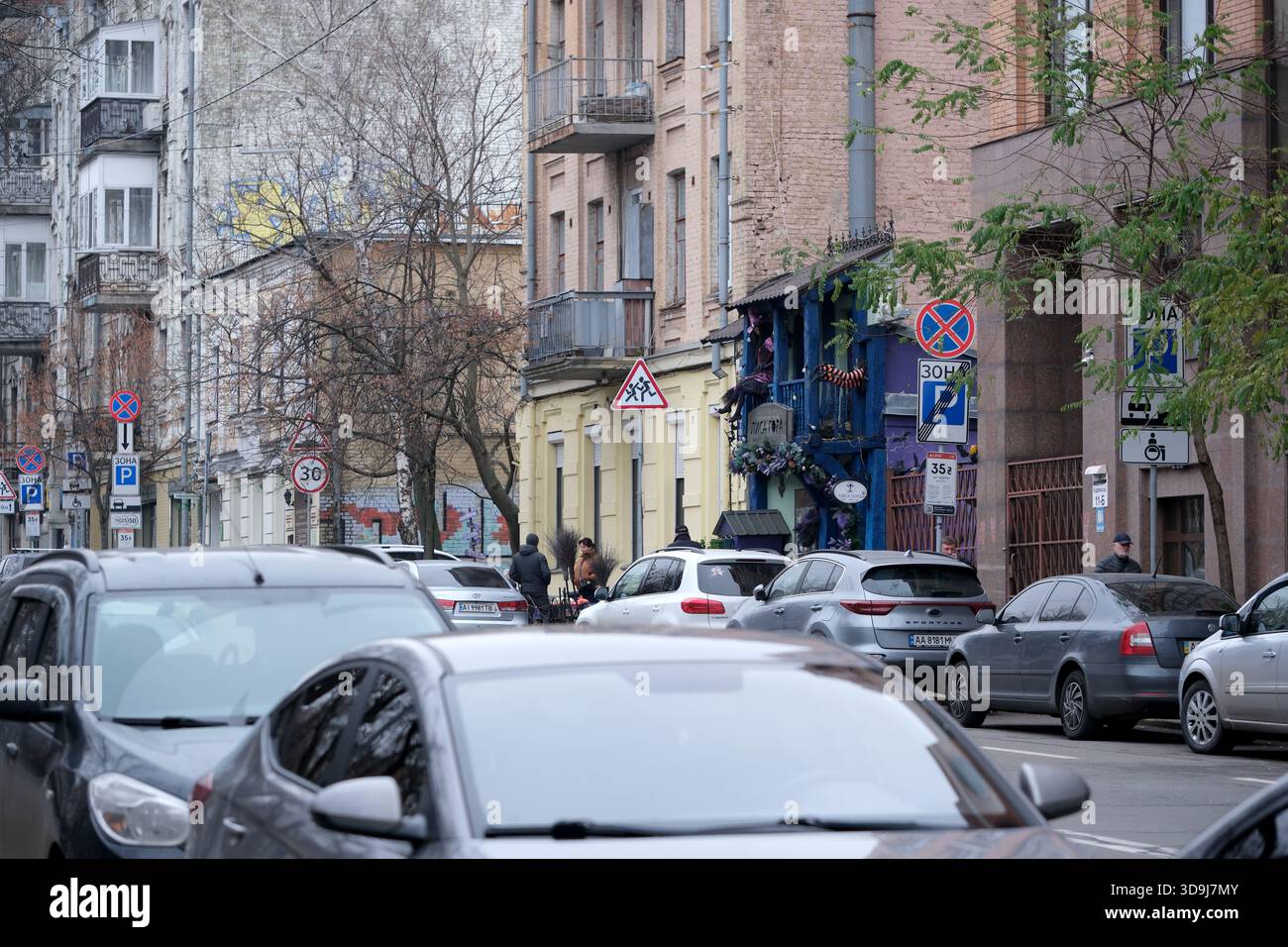 La gente cammina lungo una vivace strada costeggiata da edifici colorati e auto parcheggiate nelle vicinanze. Kiev, Ucraina. 5 dicembre 2025. Foto Stock