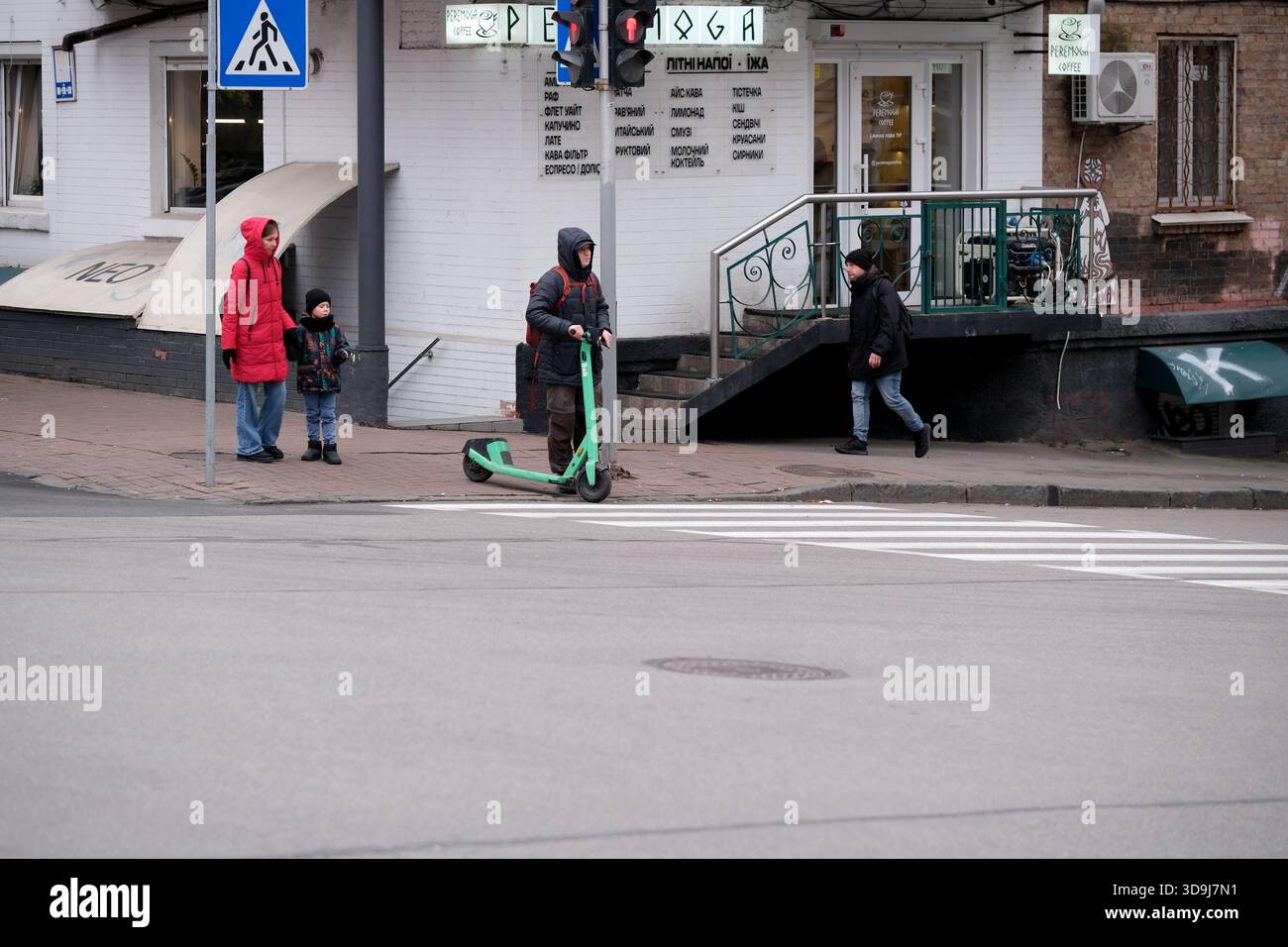 Una persona su uno scooter verde si ferma vicino a un angolo di strada affollato mentre altri passano accanto. Kiev, Ucraina. 5 dicembre 2025. Foto Stock