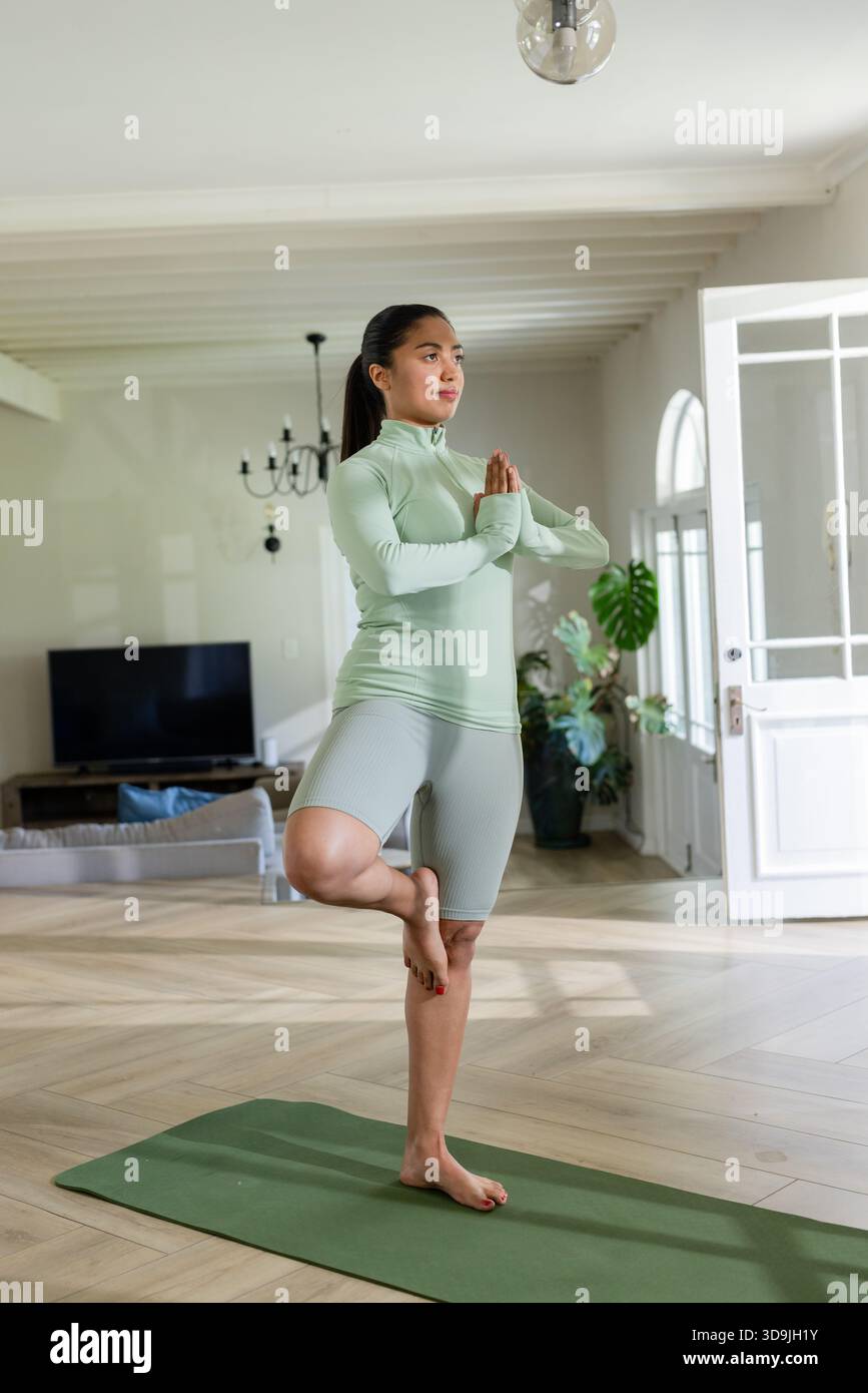 Donna asiatica in abbigliamento sportivo che tiene l'equilibrio dello yoga posa su un tappeto verde a casa con porte rivestite di vetro Foto Stock