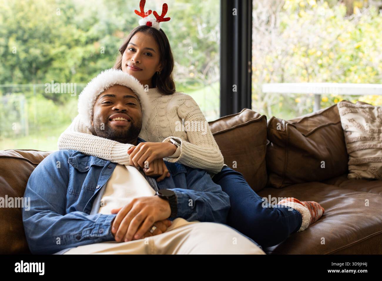 Coppia variegata che abbraccia un divano in pelle marrone da una grande finestra con fascia antler, cappello di Babbo Natale Foto Stock