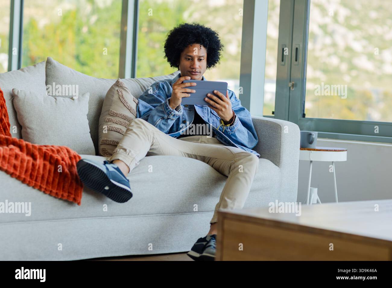Uomo afroamericano seduto su un divano grigio che regge un tablet concentrandosi sullo schermo nel moderno soggiorno Foto Stock