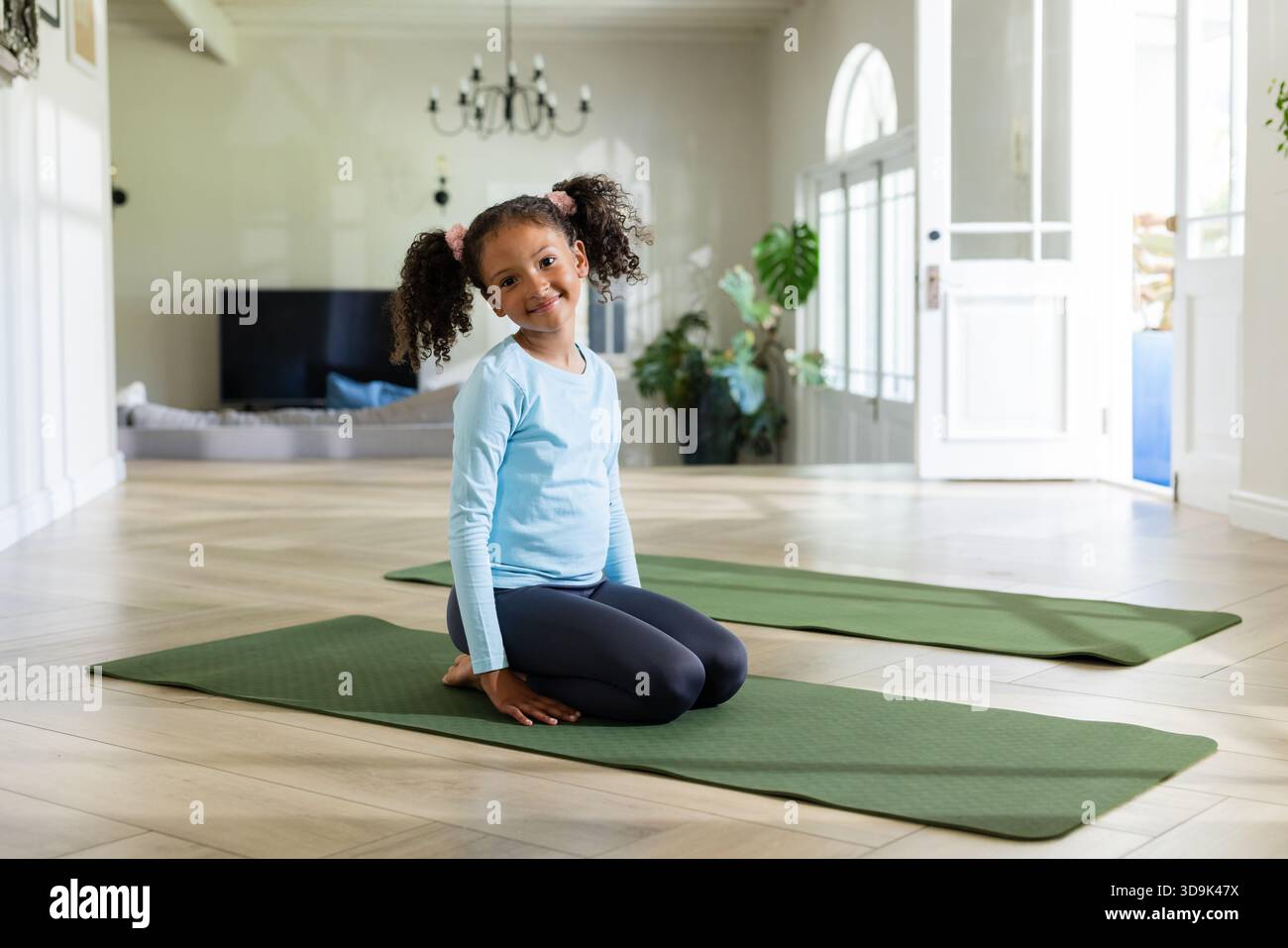 Ragazza afroamericana inginocchiata su un tappetino verde da yoga nel soggiorno accanto a porte ad arco e piante Foto Stock