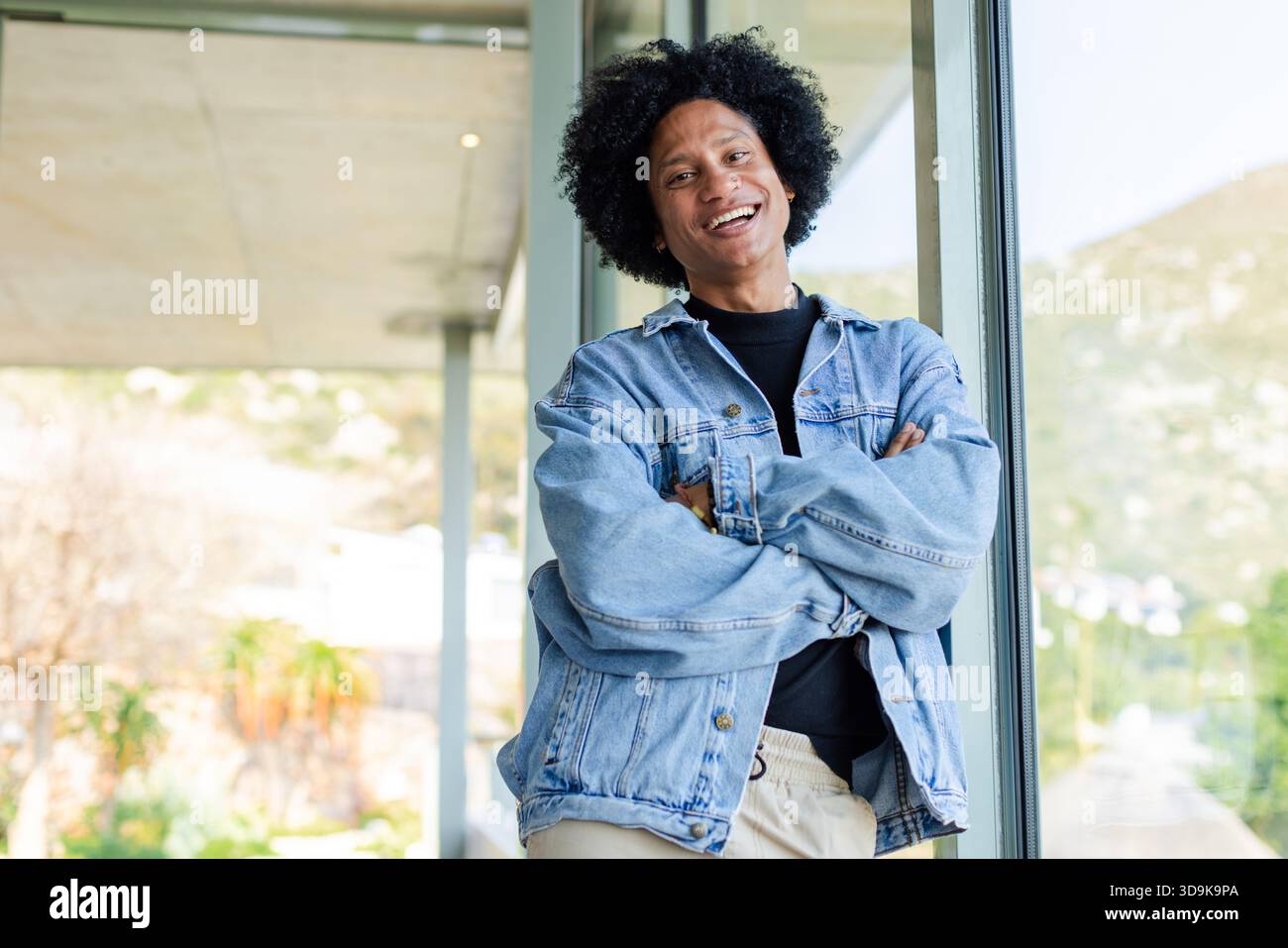 Uomo afroamericano appoggiato su una finestra di vetro all'interno di un edificio moderno sorridente in giacca di denim Foto Stock