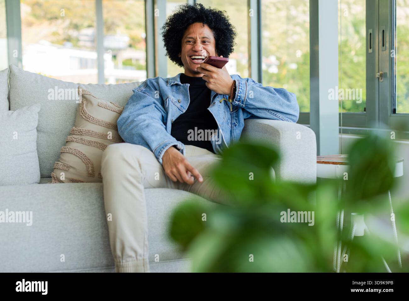 Uomo afro-americano che tiene uno smartphone color rosa e parla sul divano del soggiorno con cuscino Foto Stock