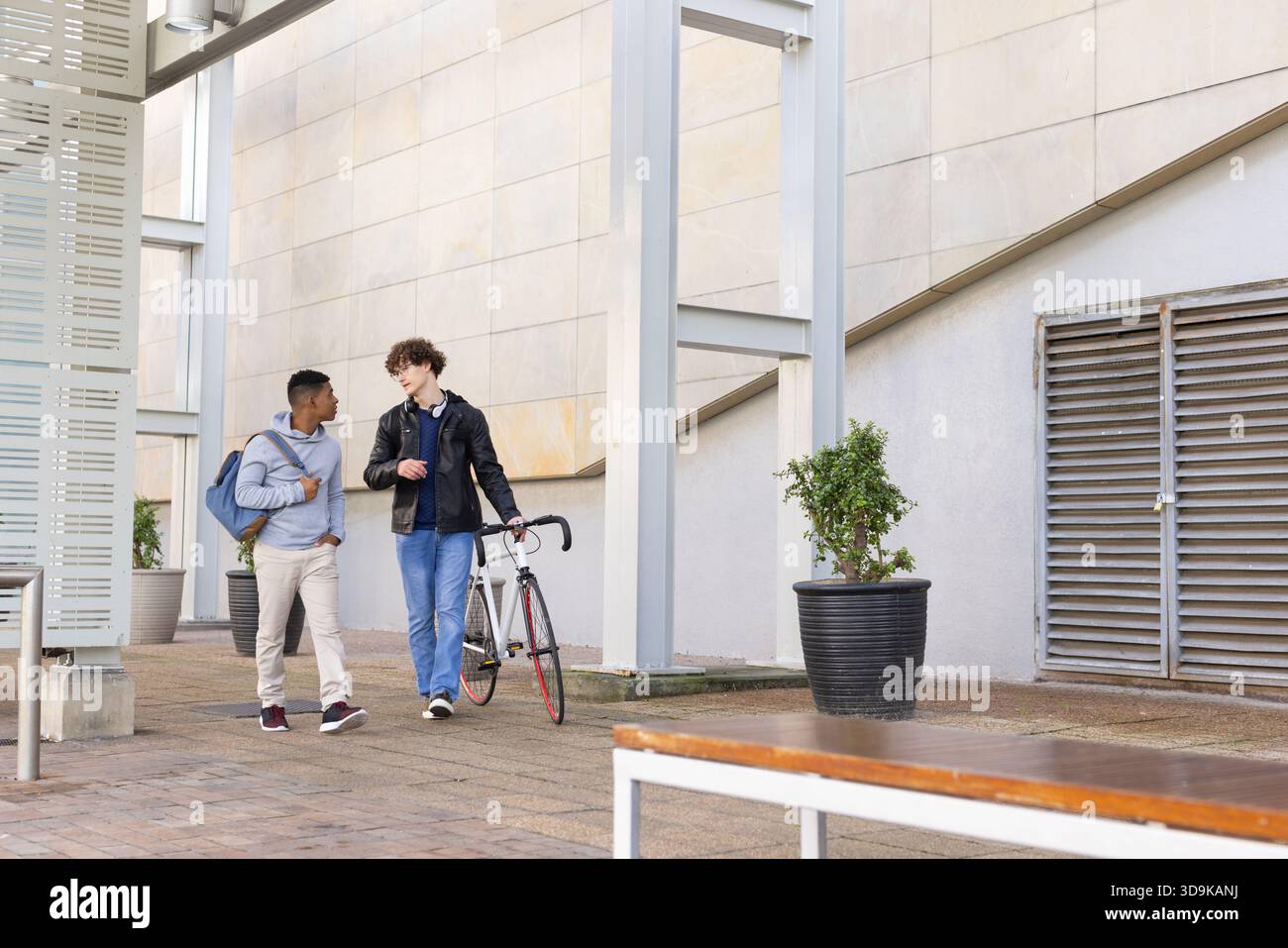 Diversi amici maschi che passeggiano lungo il percorso del campus spingono la bicicletta da strada portando zaino e cuffie Foto Stock