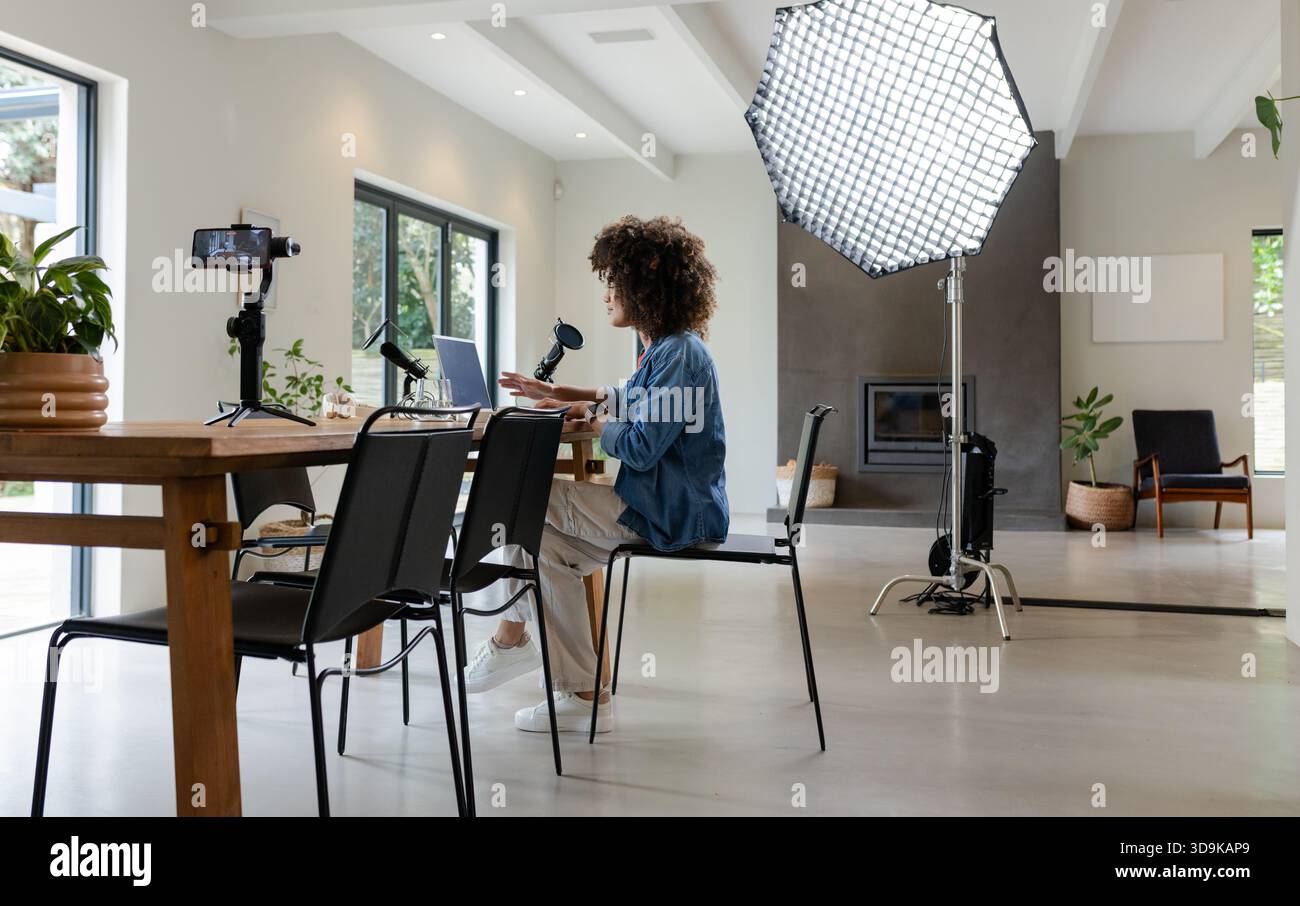 Donna seduta a un tavolo di legno che crea contenuti con il computer portatile e lo smartphone sotto la luce softbox Foto Stock