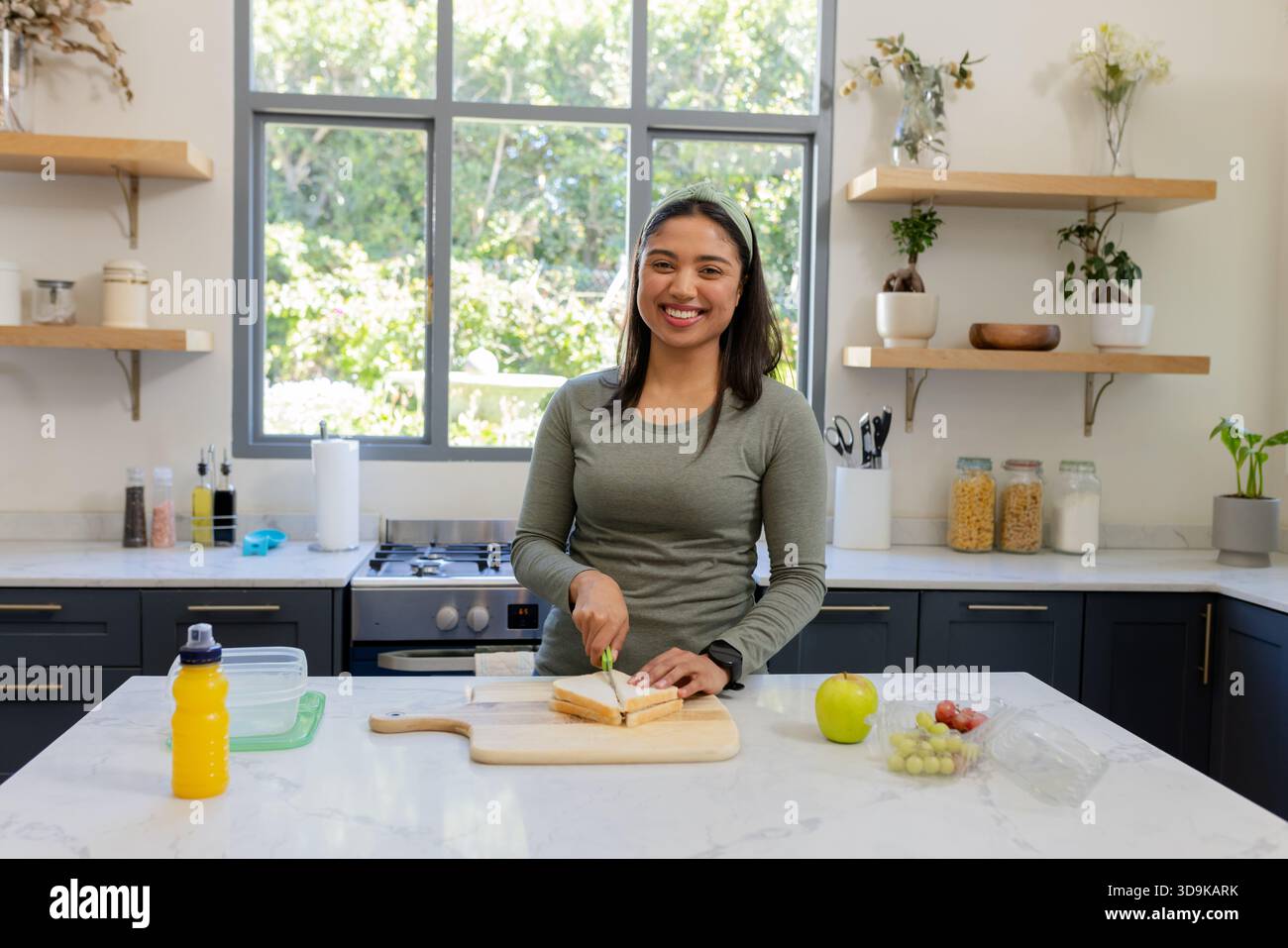 Donna che affetta panino su un piano di marmo nella cucina di casa con tagliere e contenitori di frutta Foto Stock