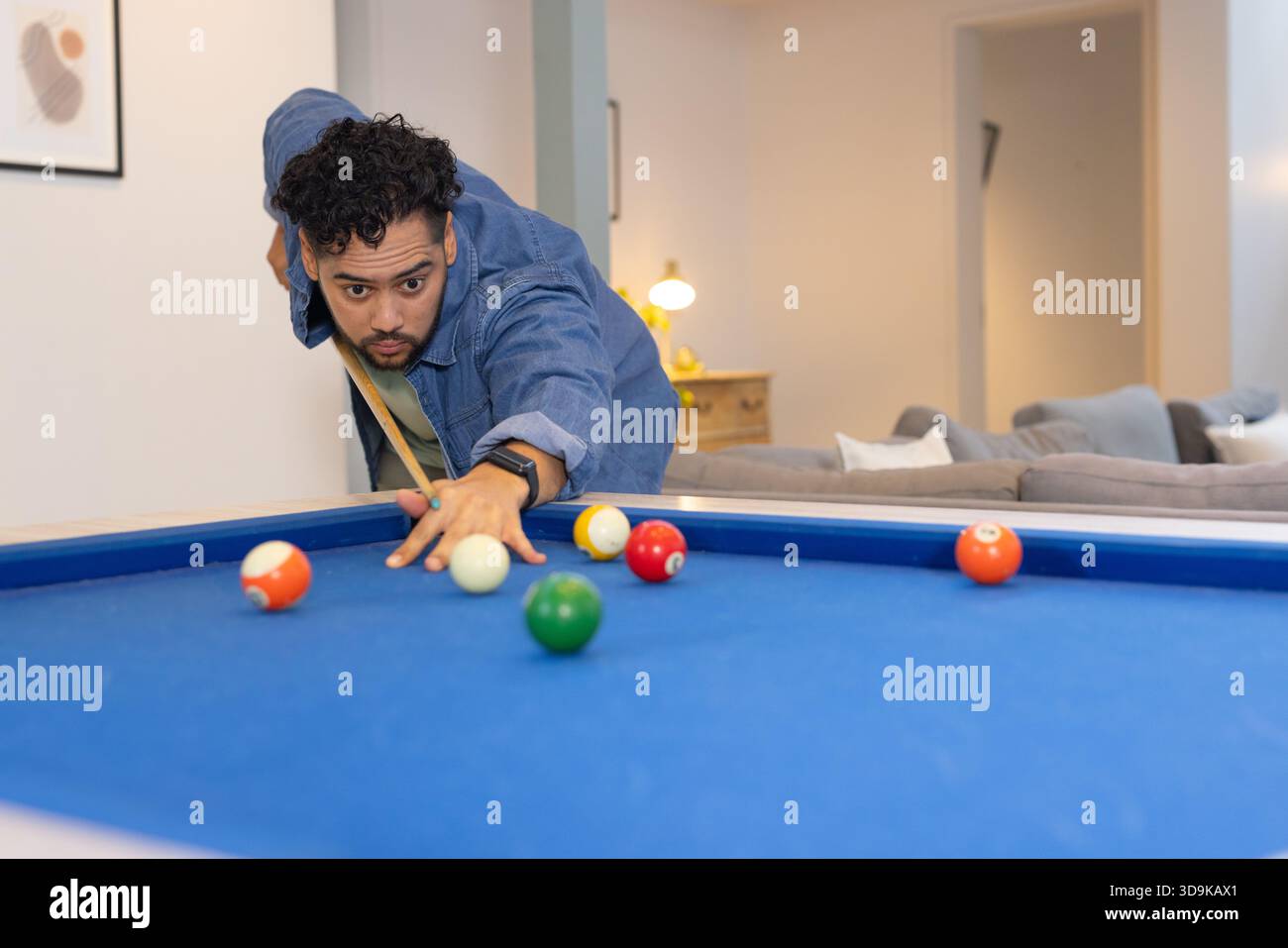 Uomo che si appoggia al tavolo da biliardo allineandosi al tiro con il bastoncino in mezzo alle palle da biliardo a casa Foto Stock