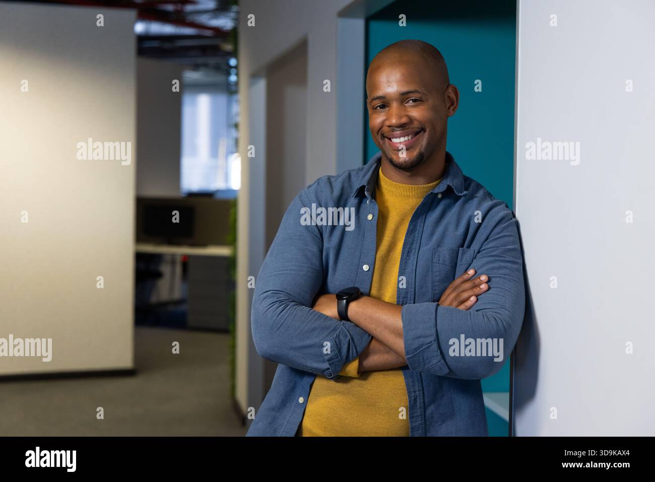 Uomo afroamericano appoggiato al muro nel corridoio dell'ufficio con smartwatch accanto al monitor del computer Foto Stock