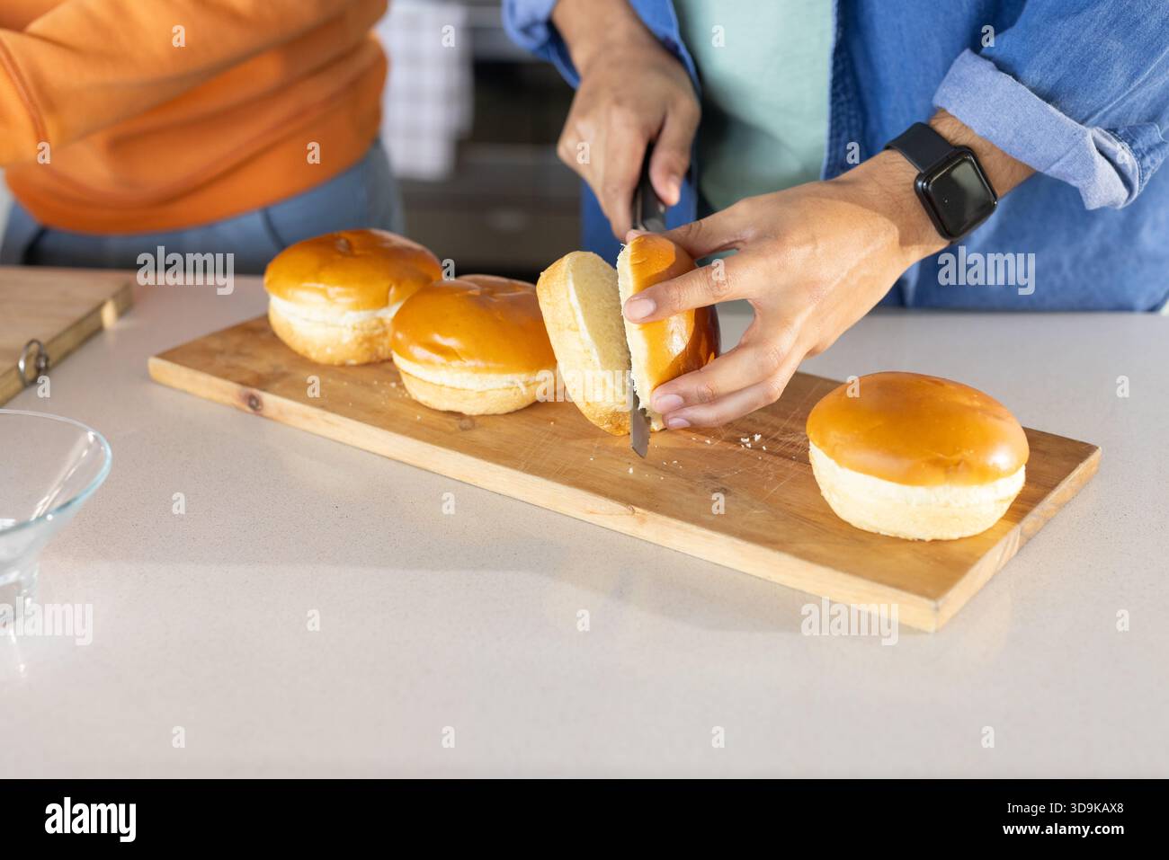 Taglio di panini brioche con coltello seghettato, amici maschi che indossano camicia in denim in cucina Foto Stock