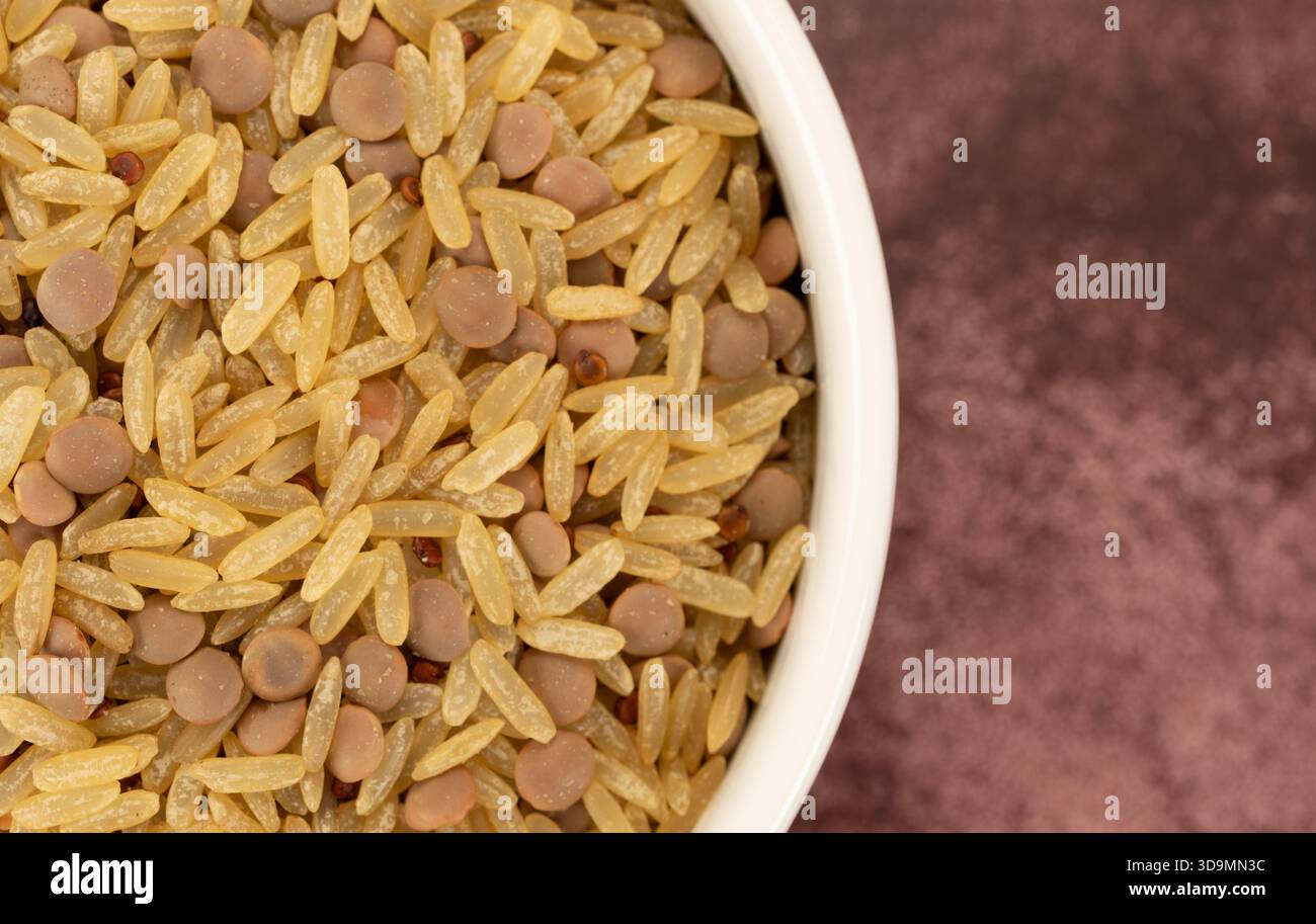Vista ravvicinata dall'alto del riso e delle lenticchie, il cibo secco si mescola in una ciotola bianca in cima a un piano portapaziente marroni strisciato Foto Stock