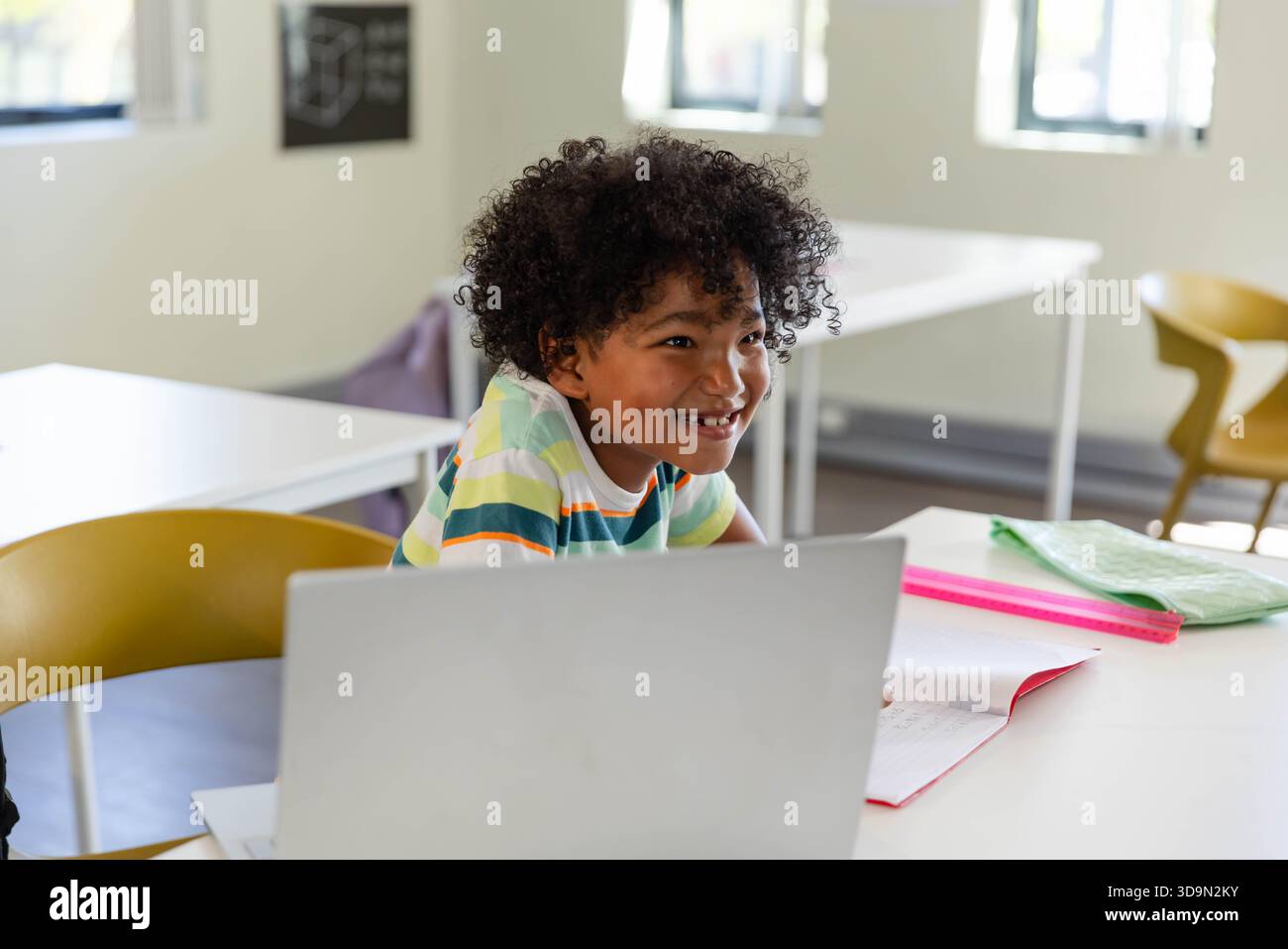 Ragazzo in una maglietta a righe sorride mentre si appoggia, utilizzando un laptop argentato al tavolo della classe con un notebook rosa Foto Stock