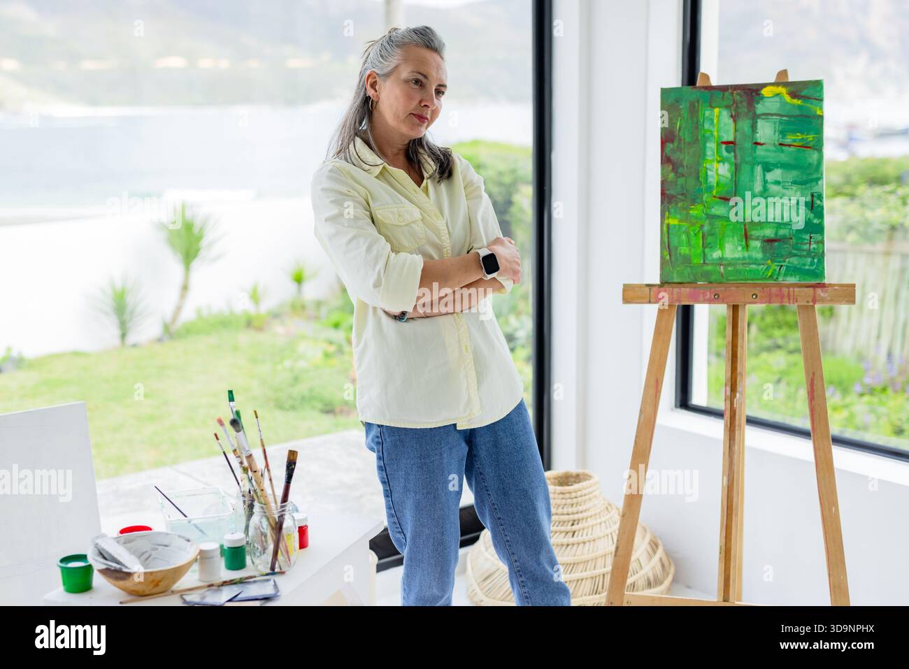 Donna anziana in piedi accanto a un cavalletto di legno che tiene la pittura astratta verde in uno studio d'arte con pennelli Foto Stock
