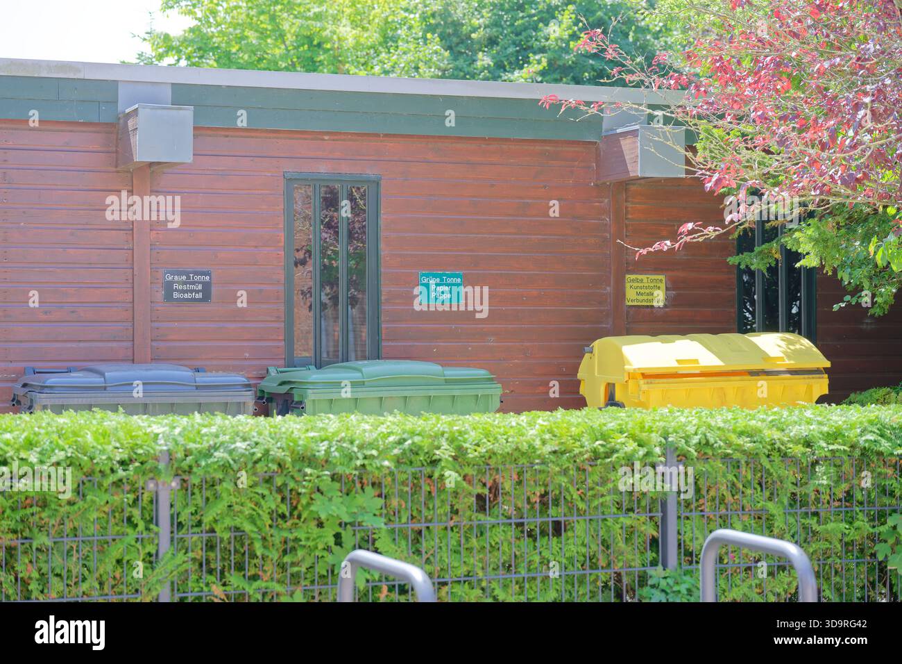 I contenitori color Code gialli, verdi e grigi si trovano dietro una recinzione vicino a un edificio in legno, circondati dal verde rifinito in una giornata di sole Foto Stock