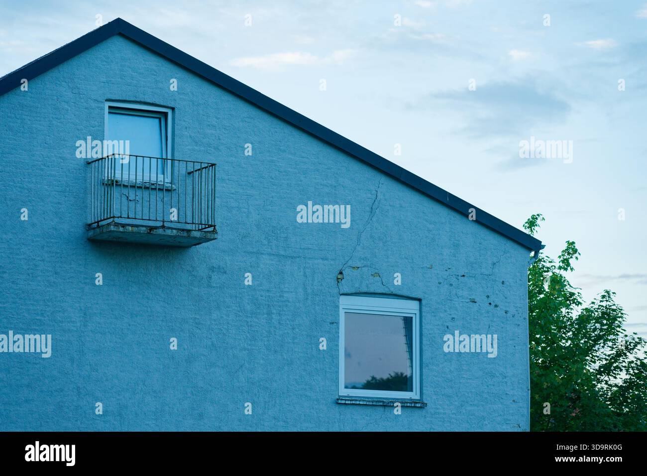 Una casa bianca mostra una parete laterale sotto il tetto con un balcone attaccato ad una finestra senza porta, con una piccola finestra di plastica sotto e un avviso Foto Stock