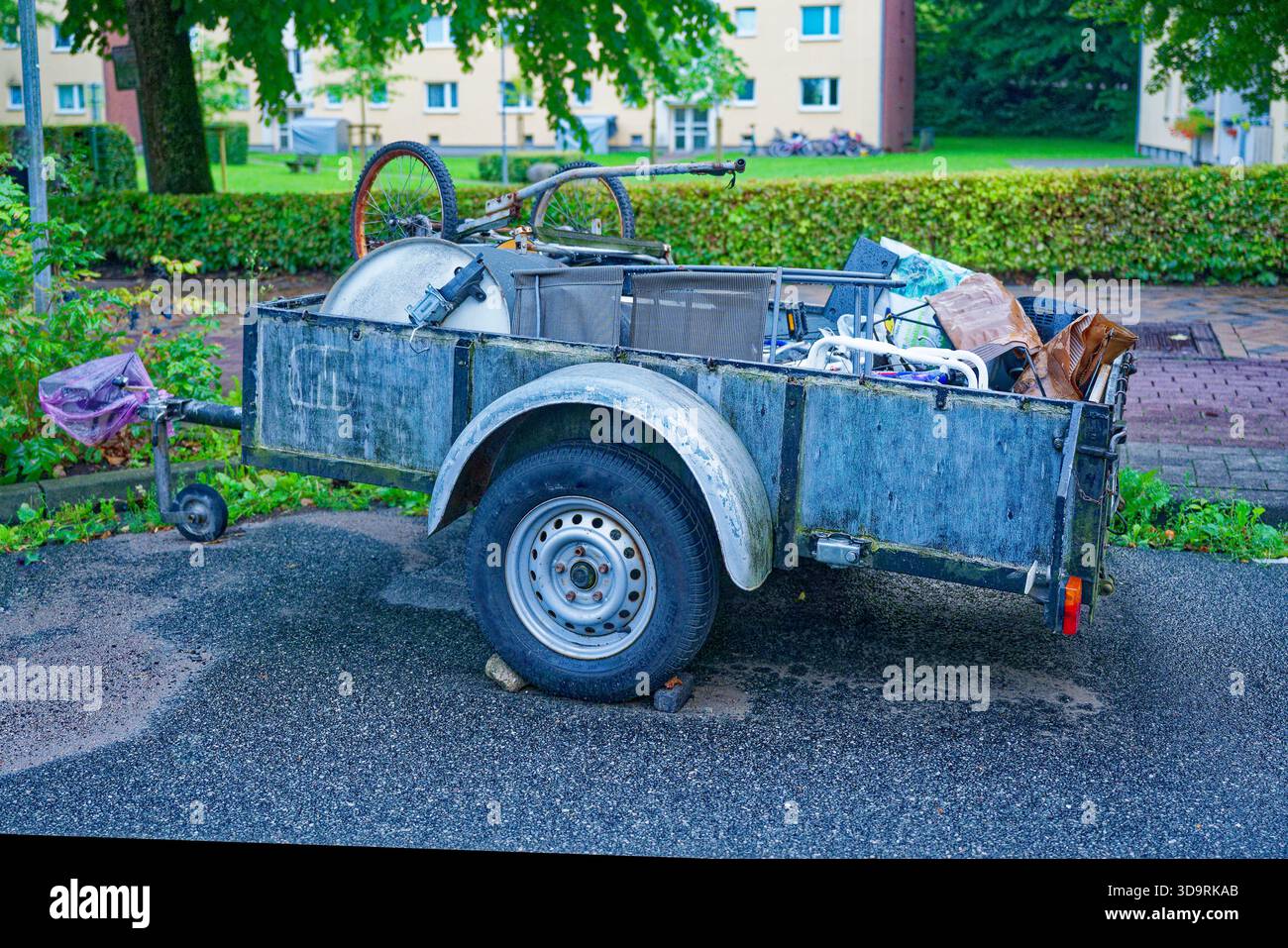 Un vecchio rimorchio a due ruote è parcheggiato con pietre sotto le ruote. È carico di vari oggetti scartati, come una bicicletta rotta, una sedia e un piatto, Foto Stock